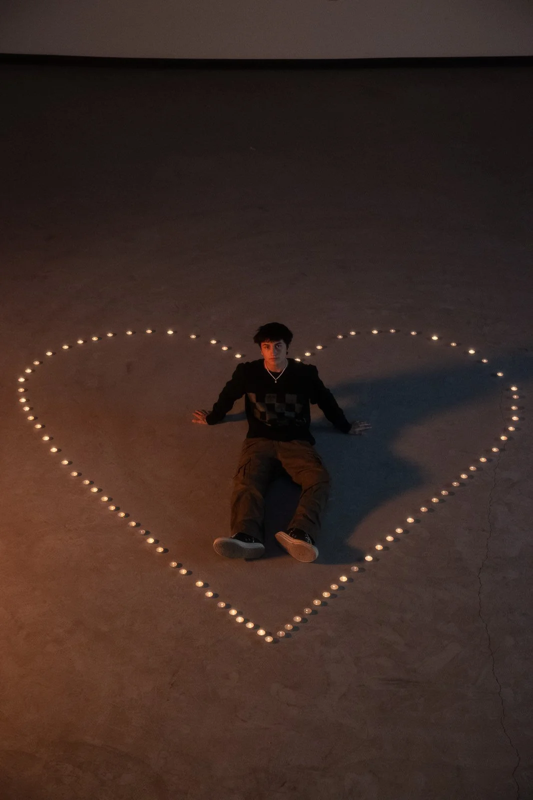 A person sitting inside a large heart shape made of small lights on the floor in a dark room.