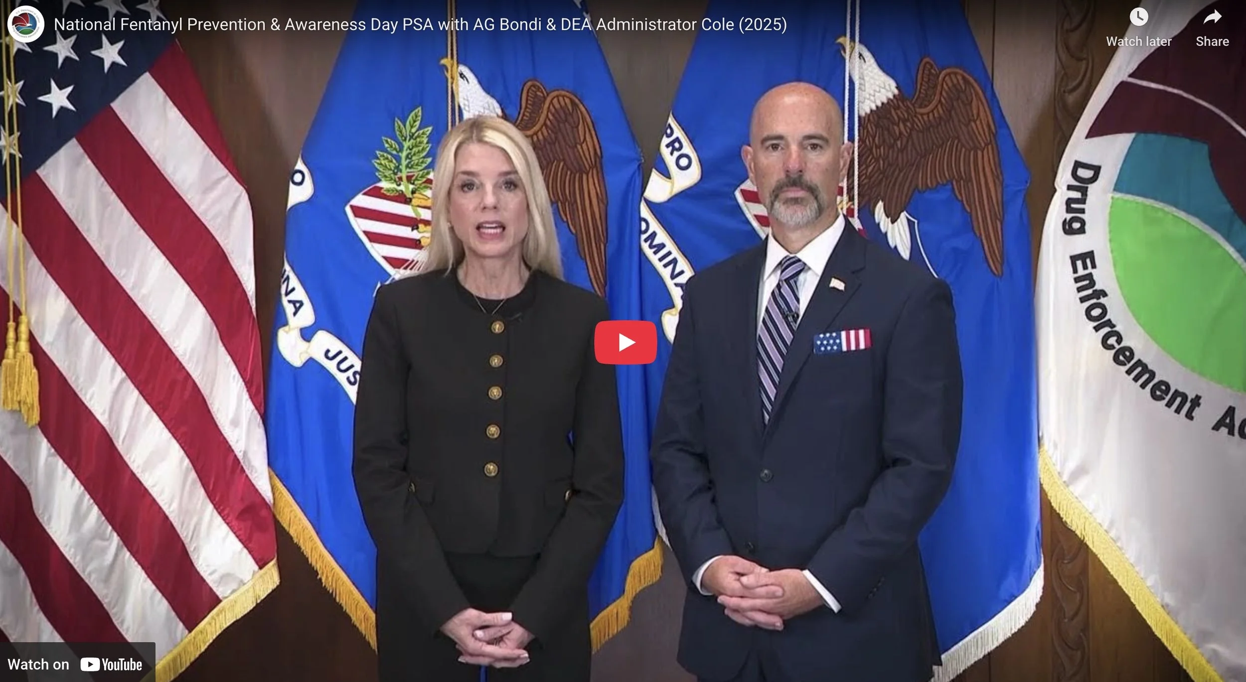 A woman and a man standing in front of multiple flags, including the American flag, the Missouri state flag, and the Department of Emergency Management flag. The woman is speaking, and both are dressed in formal attire.