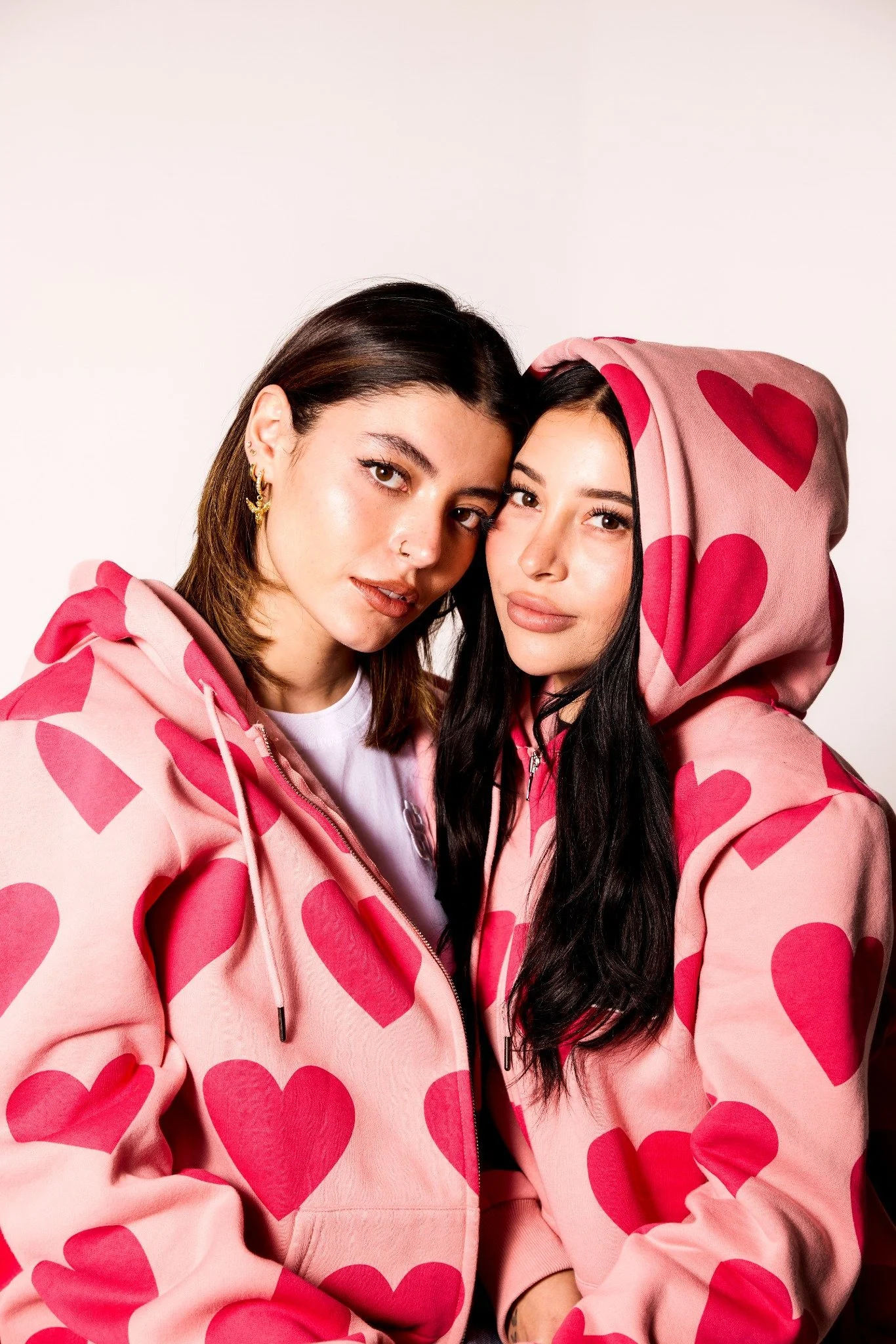 Two women wearing matching pink hoodies with red hearts, posing closely together against a plain white background.