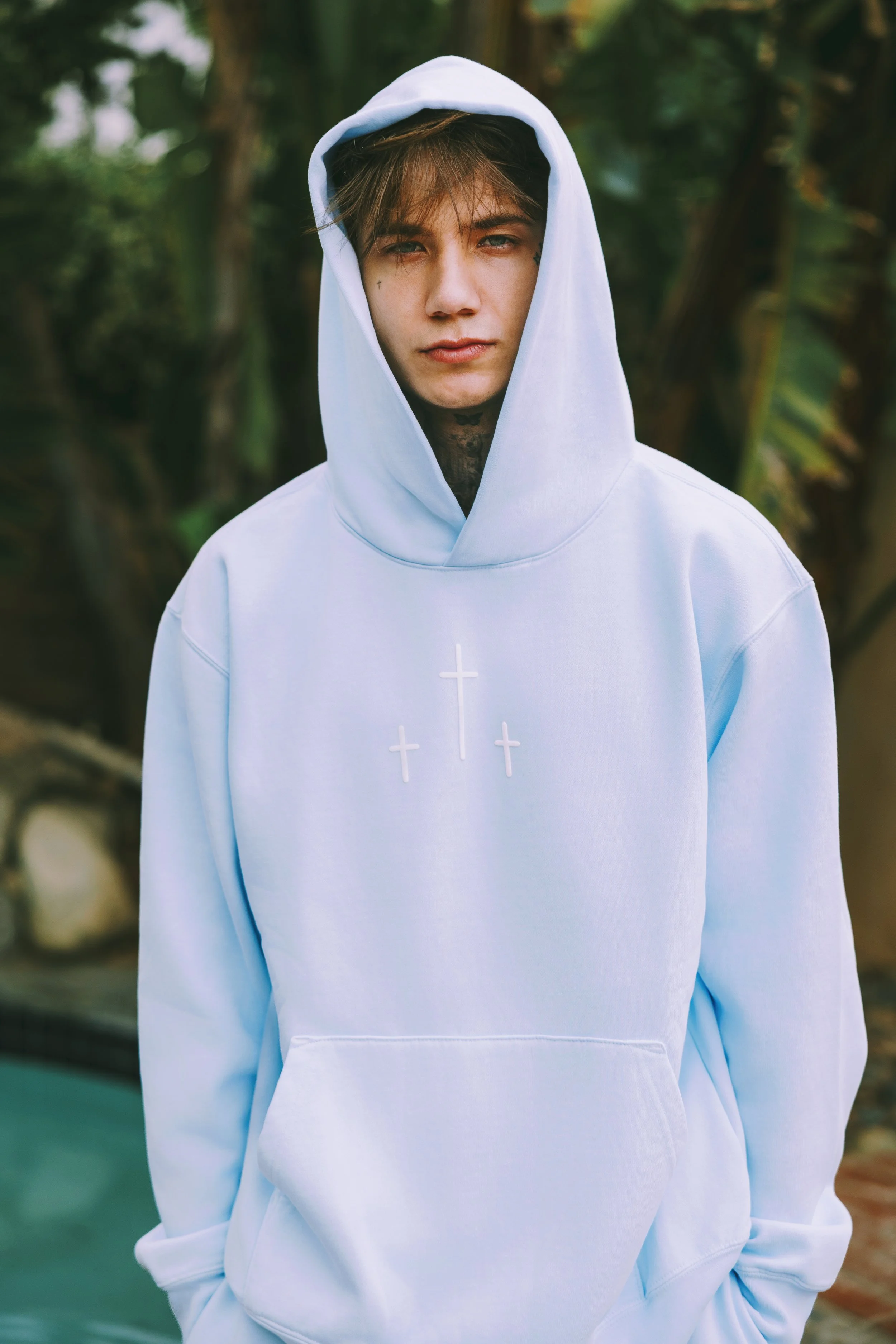 A young man with light brown hair and blue eyes wearing a white hoodie with crosses embroidered on the chest, standing outdoors with a background of green trees.