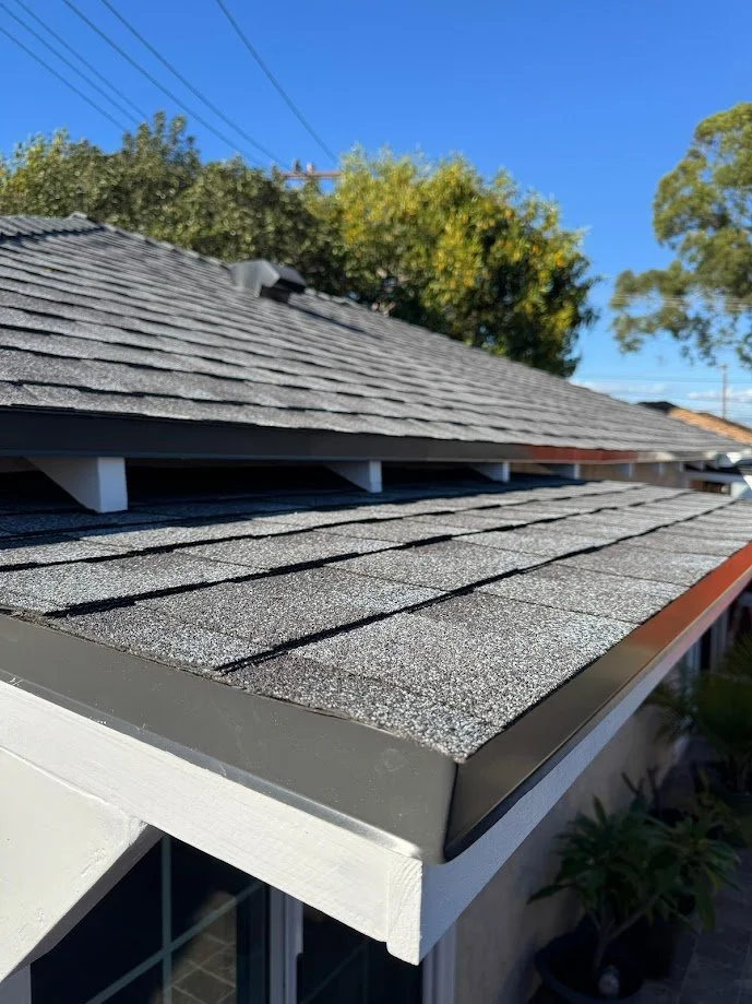 The image shows a close-up view of a house's roof with gray asphalt shingles under a clear blue sky, with trees and power lines in the background.