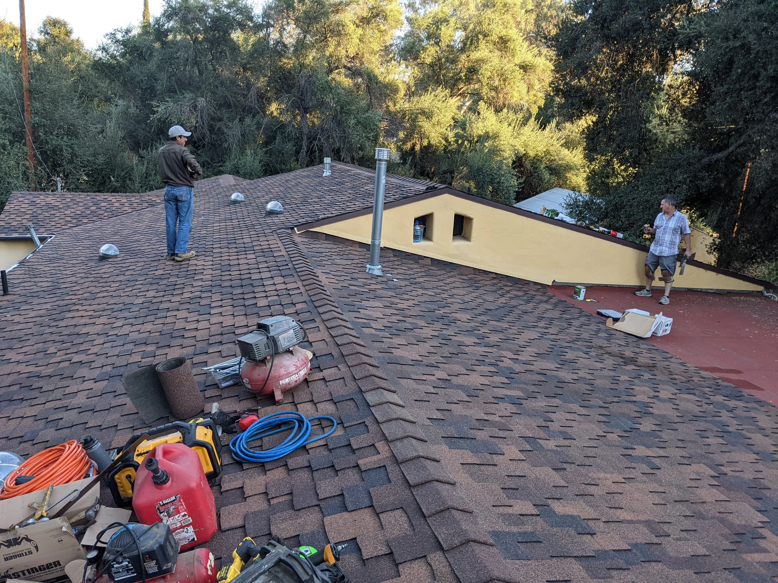 Two workers on a residential roof surrounded by tools and equipment, installing or repairing the roof under a sunny sky with trees in the background.