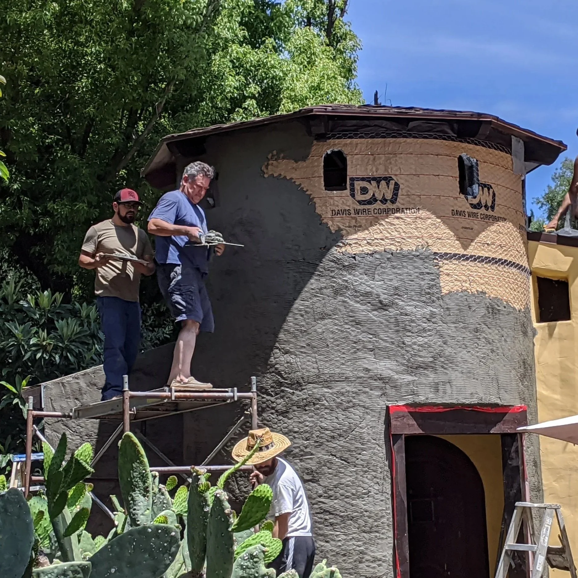 Two men standing on scaffolding working on a house built to look like a cactus with its textured surface and green cactus plants in the foreground.