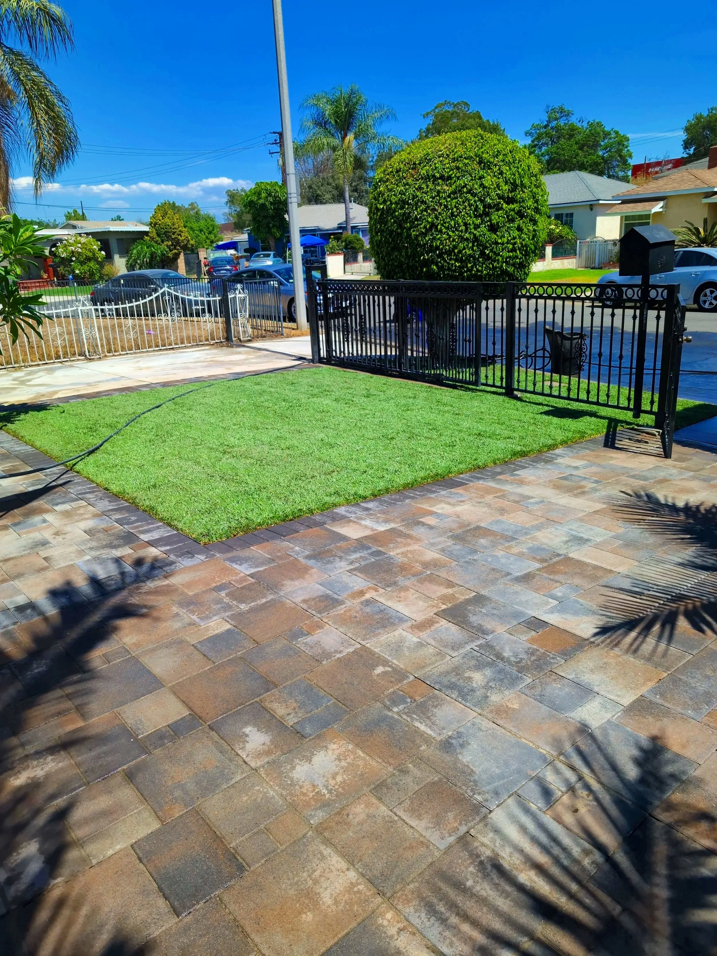 A residential yard with a small artificial grass lawn, a black metal fence gate, and a paved brick patio. There are trees and houses visible across the street under a blue sky.