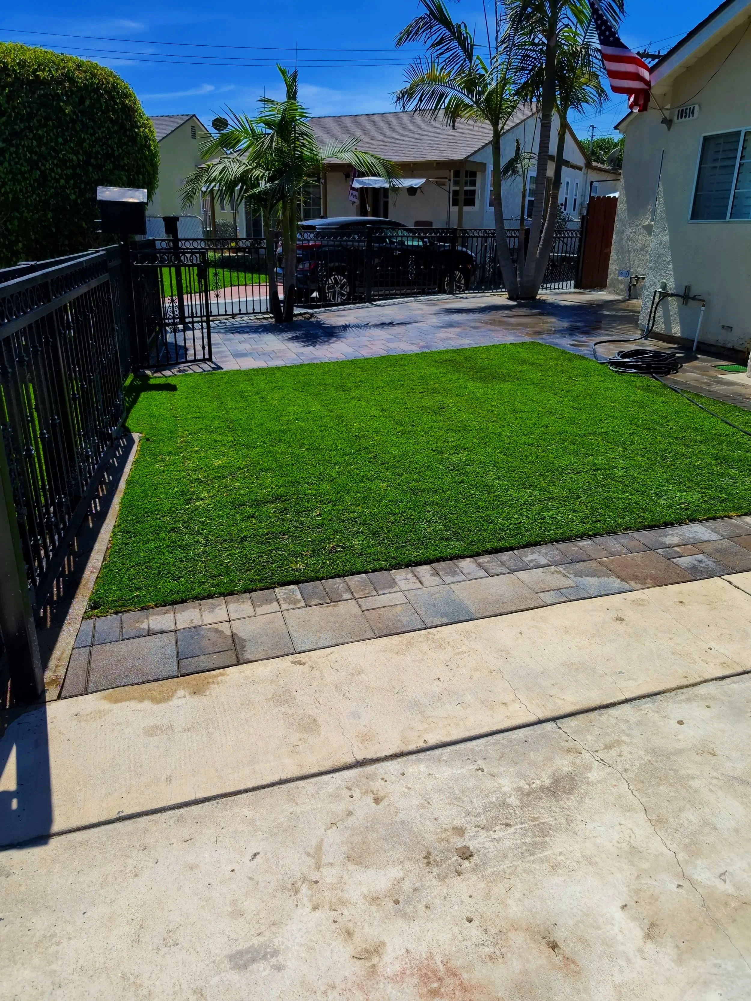 A backyard with a small patch of vibrant green grass, surrounded by tile and concrete surfaces. There is a black metal fence on two sides, with a gate on the left. In the background, there are tall palm trees, a house with a porch, and parked cars. A