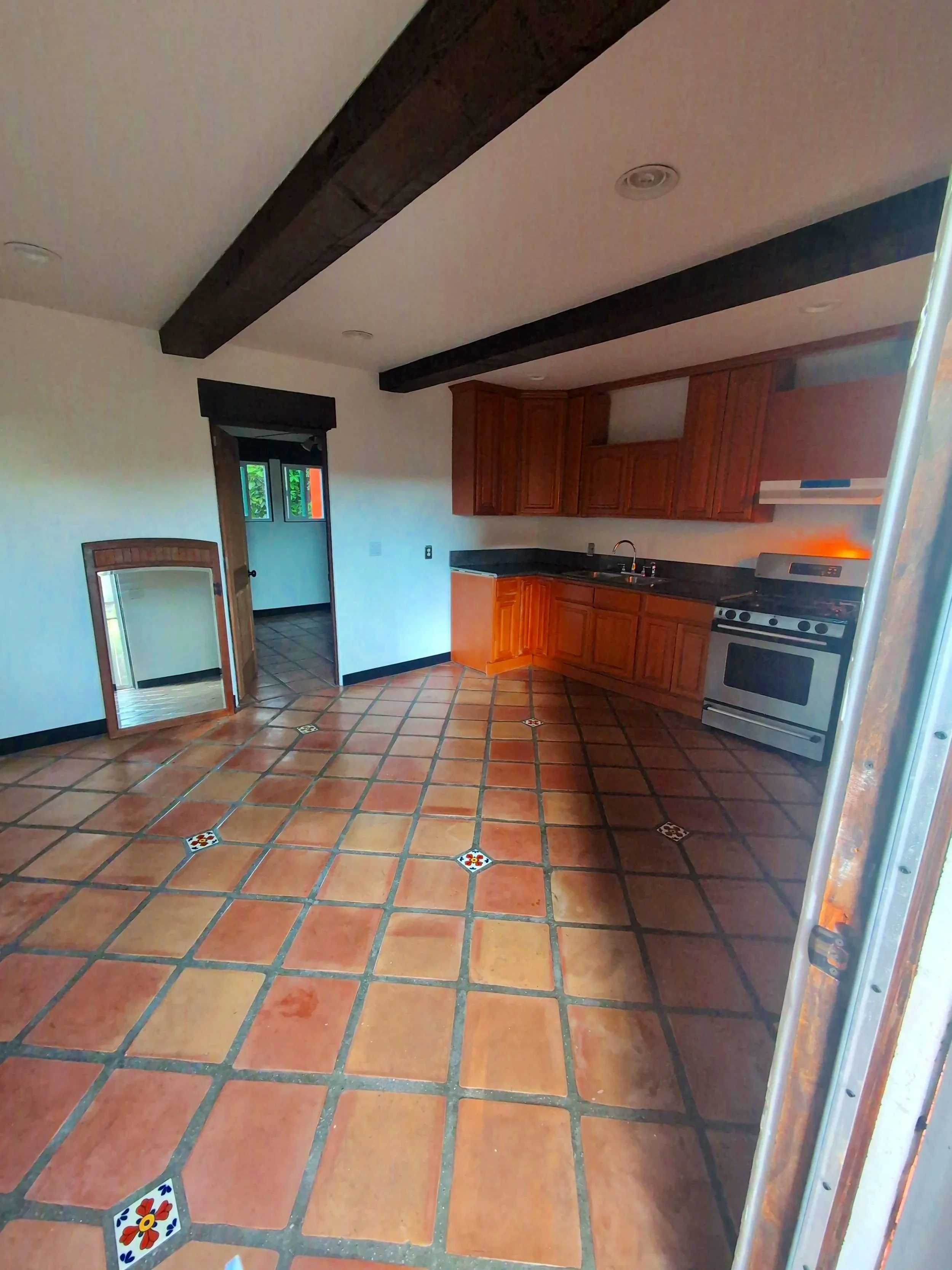 Empty kitchen with terracotta tiled floor, wooden cabinets, black countertops, and a white stove. Beams on ceiling and a mirror on the left side.
