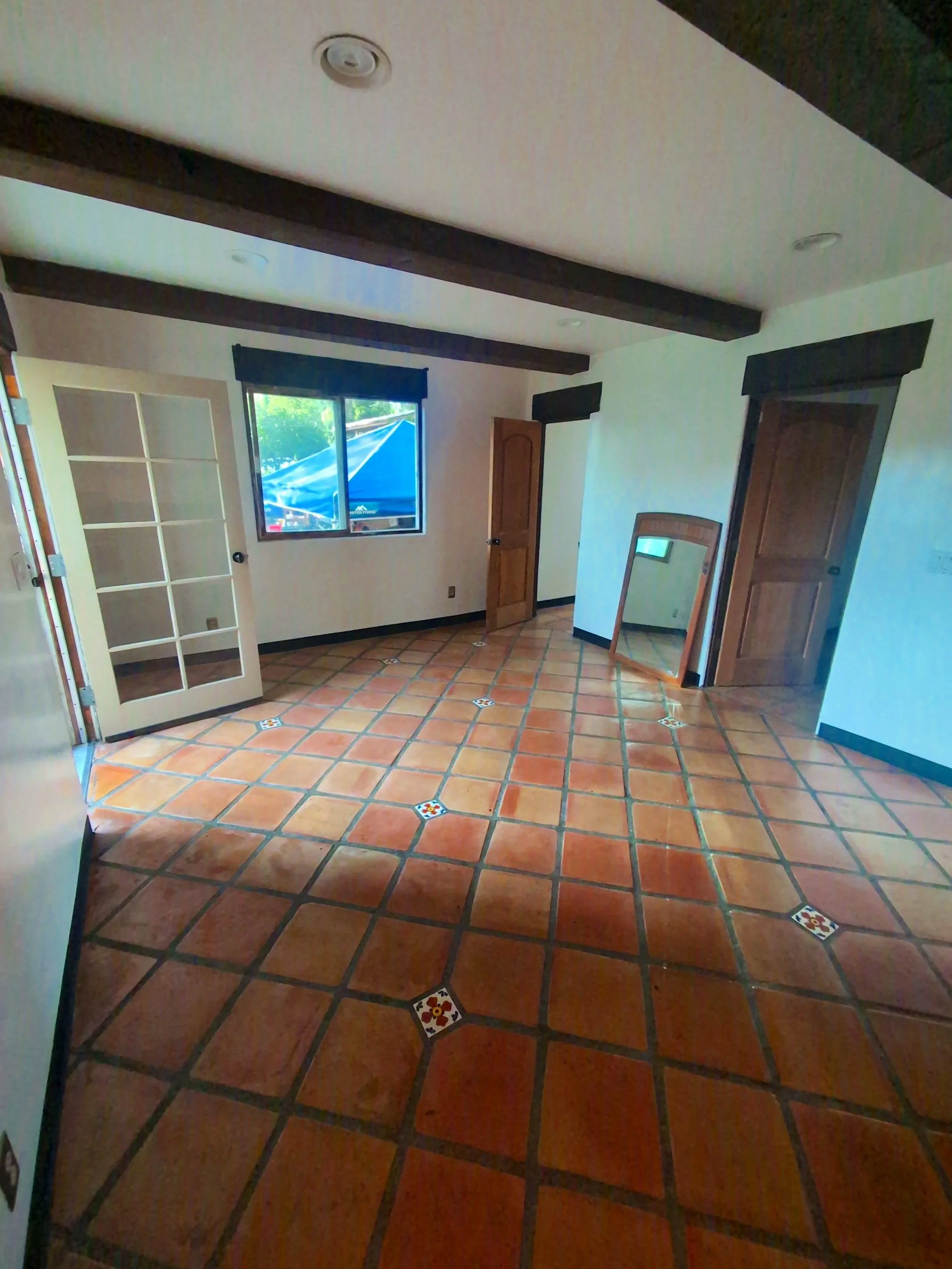 Empty room with terracotta tile floor, a large window, a wooden door, a mirror leaning against the wall, and a glass-paned door with black trim. The ceiling has dark wooden beams.