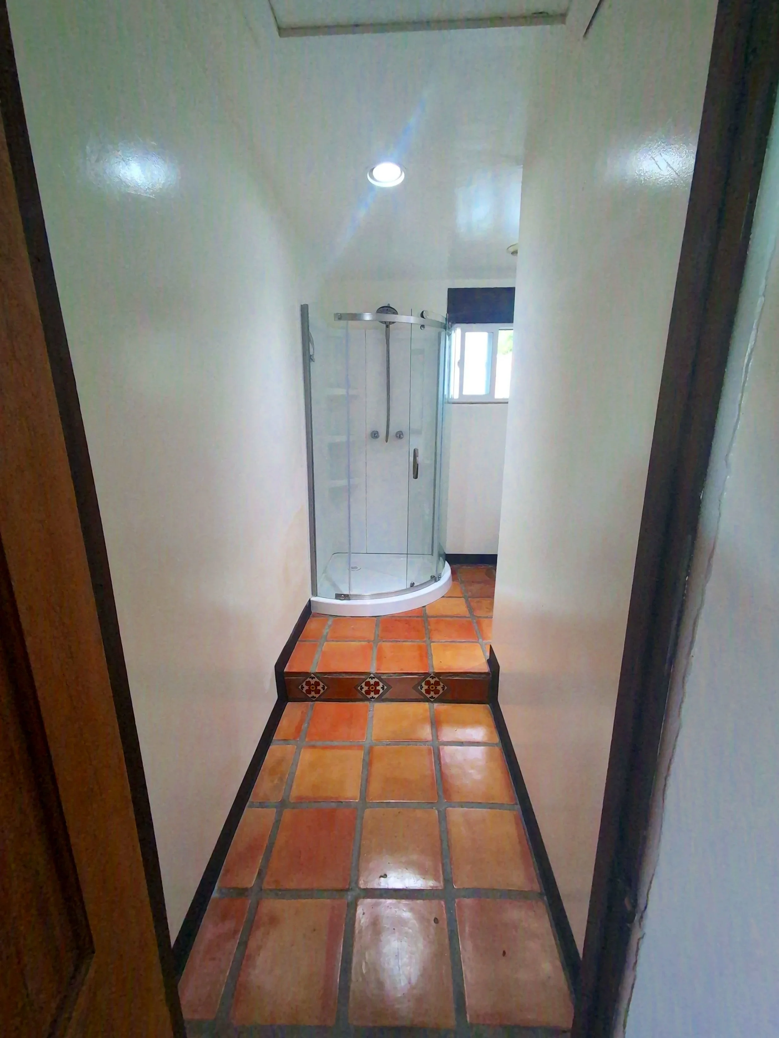 A narrow hallway with terracotta tiled floor leading to a corner with a small shower stall next to a window.