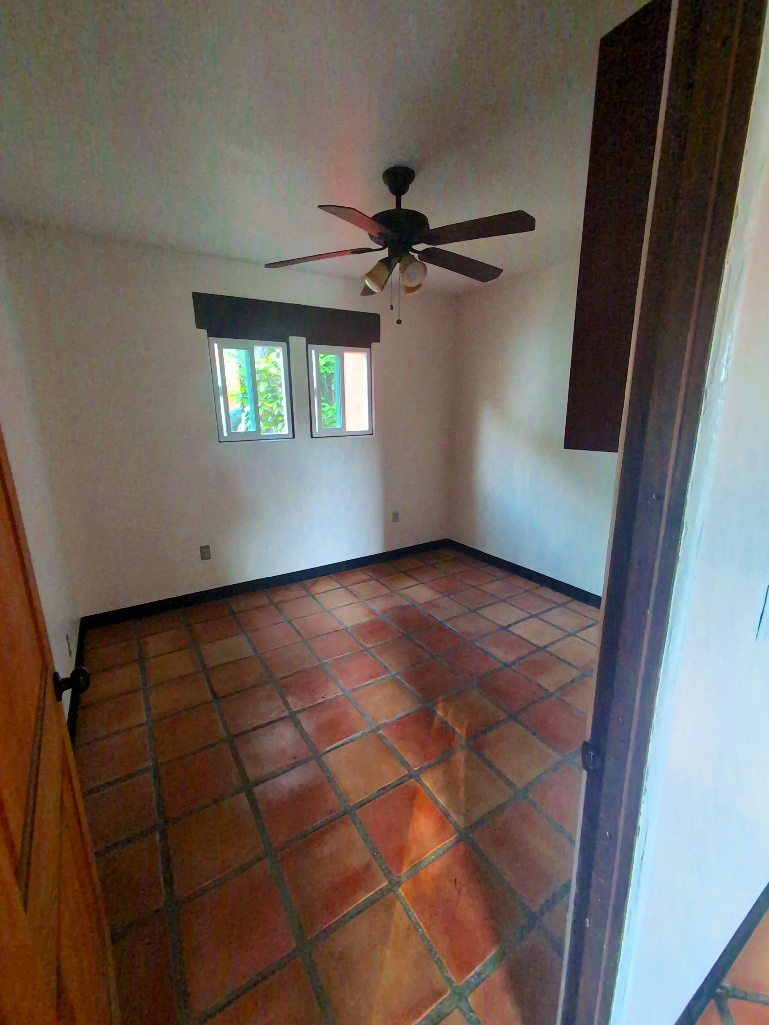 Empty room with terracotta tile flooring, white walls, three small windows with black trim, a dark wood closet door, and a ceiling fan with four blades and attached light fixtures.