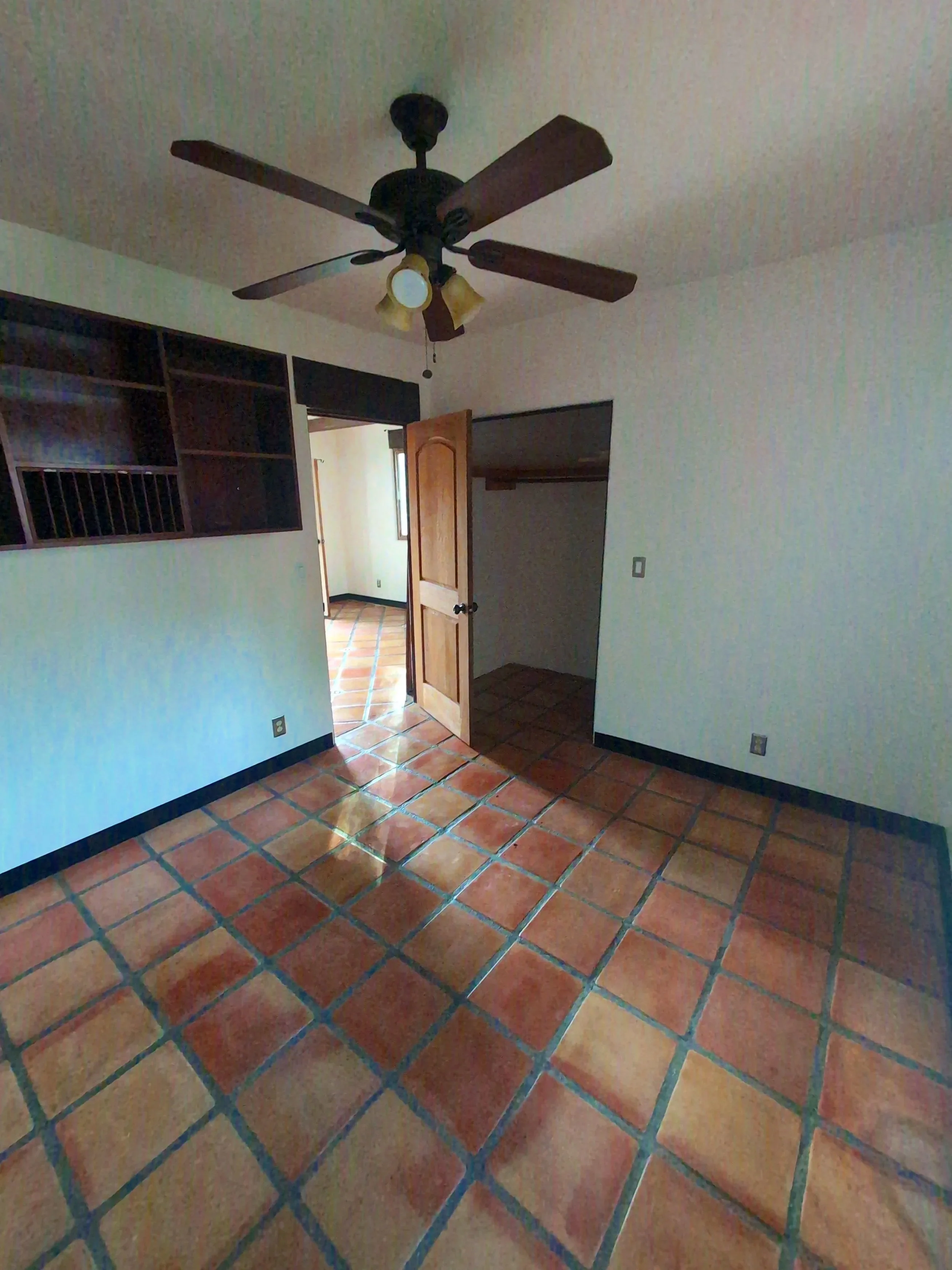 Empty room with terracotta tile flooring, a ceiling fan with lights, an open closet, and a doorway leading to another room with similar flooring.