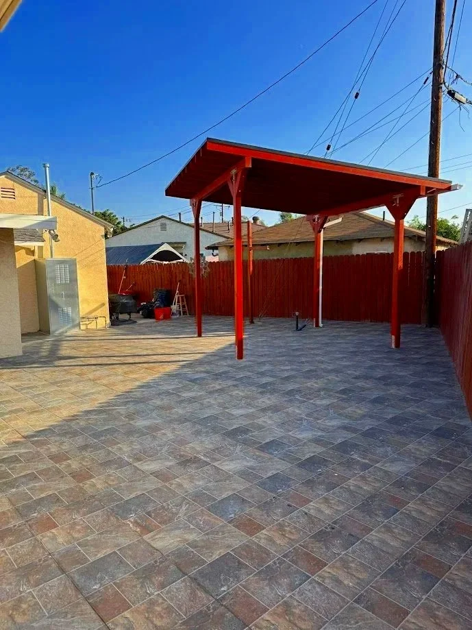 A backyard with a paved patio, a red wooden gazebo frame, and a red fence in the background under a clear blue sky.