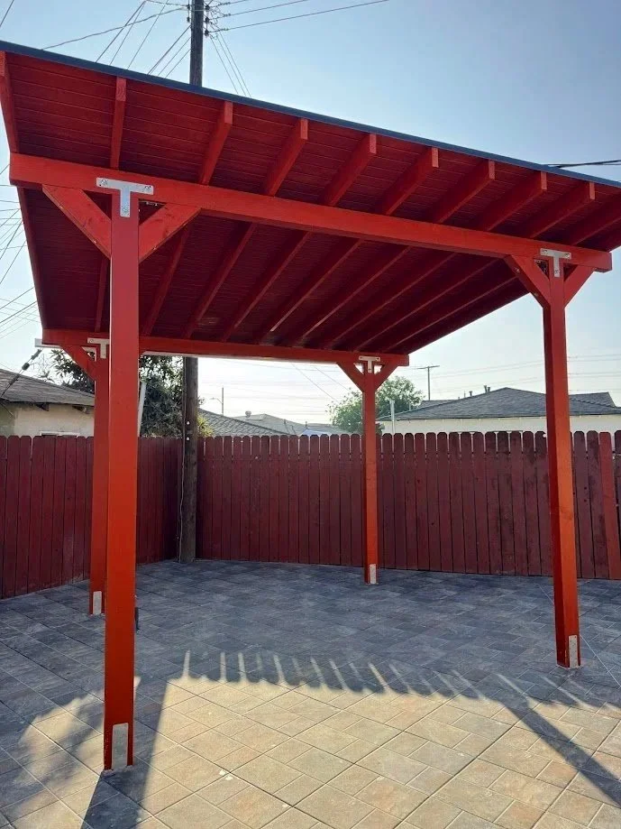 A red wooden carport with four posts, located on a paved patio, with a red wooden fence and neighborhood houses in the background.