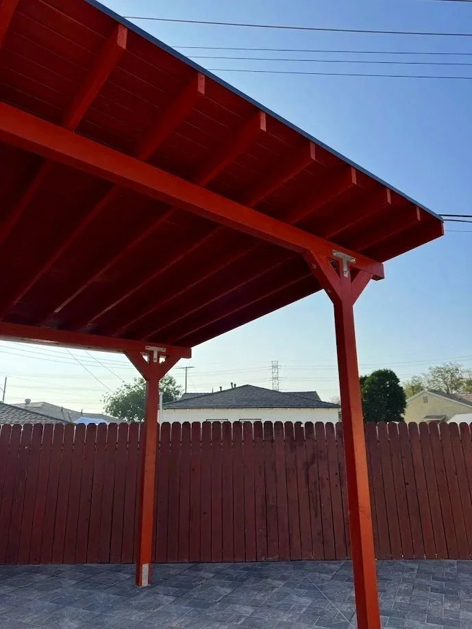 A red wooden patio cover with support beams and a tiled floor, enclosed by a red wooden fence and clear blue sky in the background.