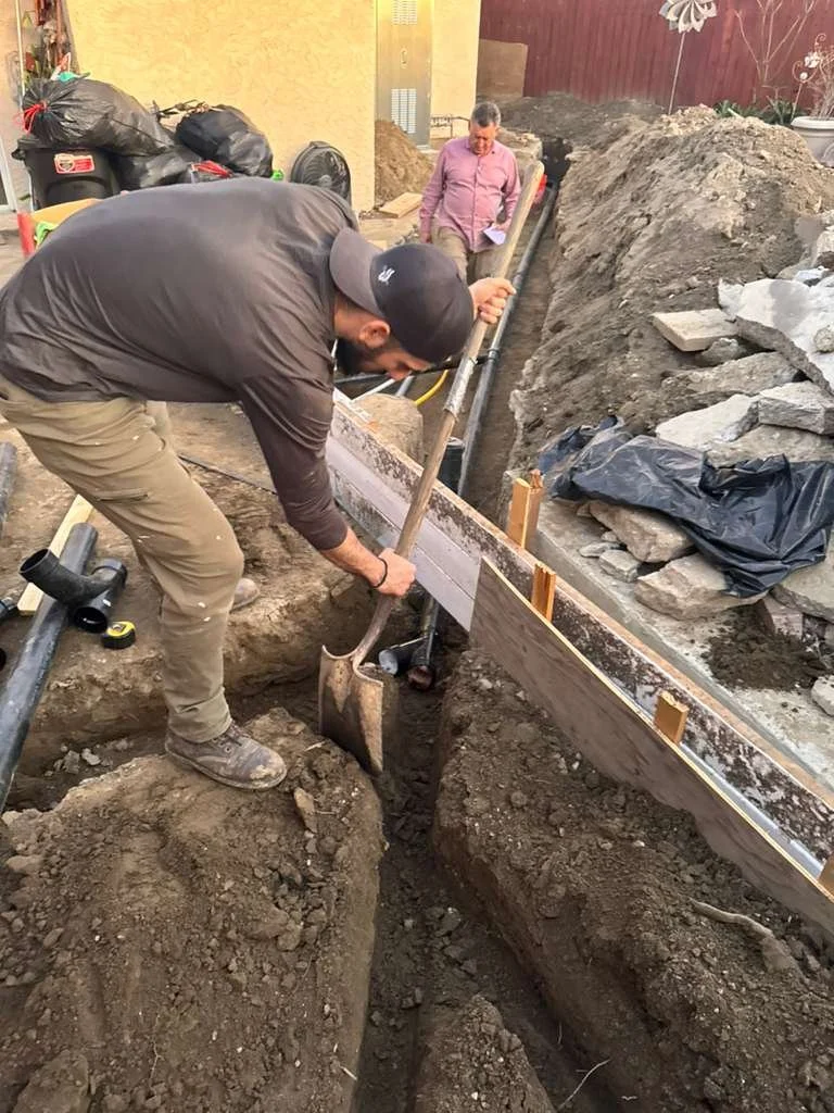 Man digging a trench with a shovel while another person observes in the background.