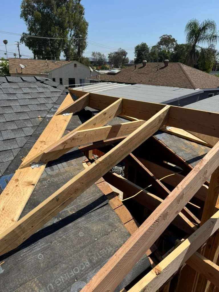 Construction of a roof with exposed wooden framing and roof sheathing in progress, residential neighborhood in the background.