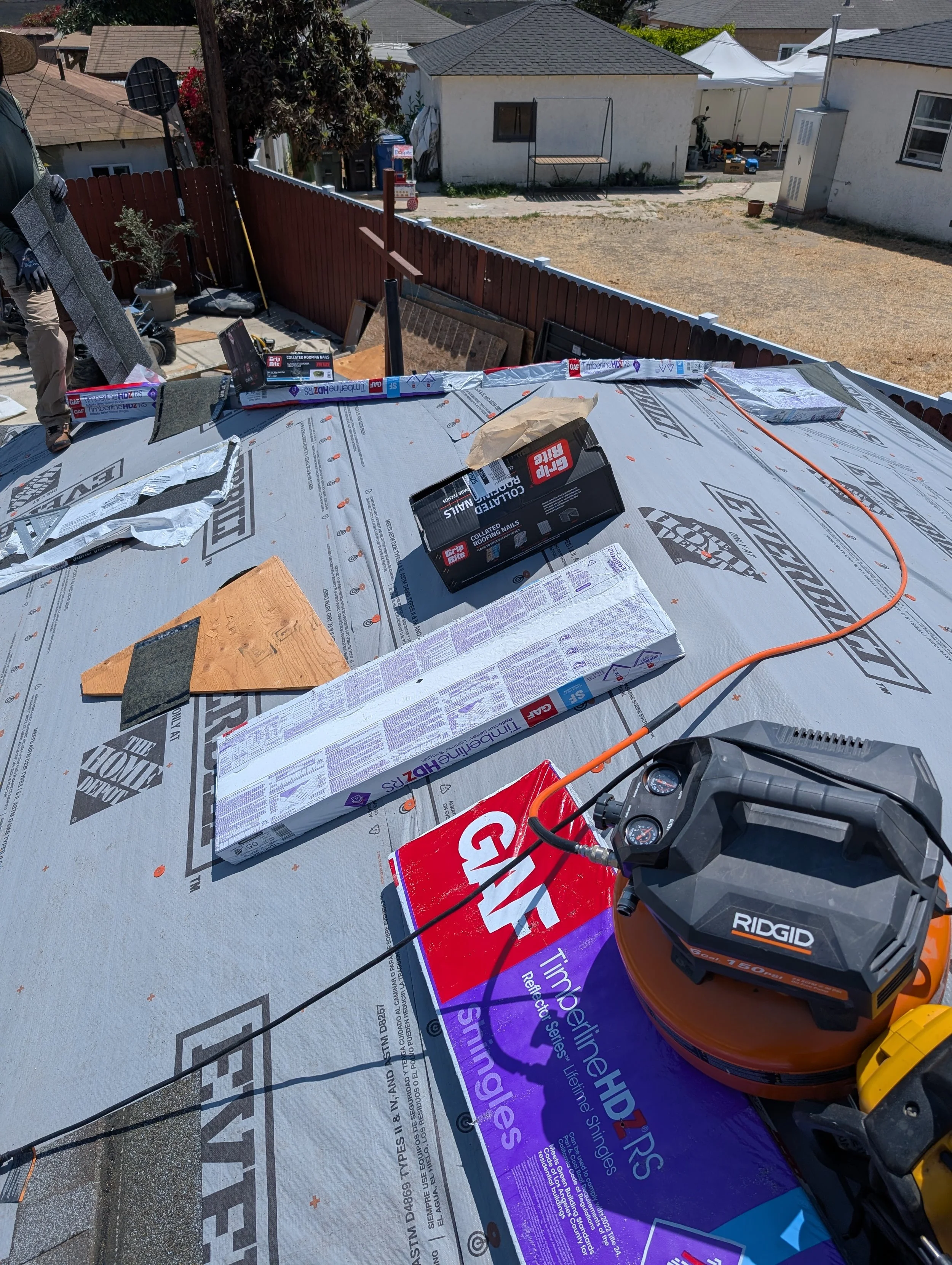 Construction site on a house roof with roofing materials, tools, and equipment visible, including a Ridgid air compressor, roofing underlayment, and boxes of roofing nails or screws, with neighboring houses and a fenced backyard in the background.