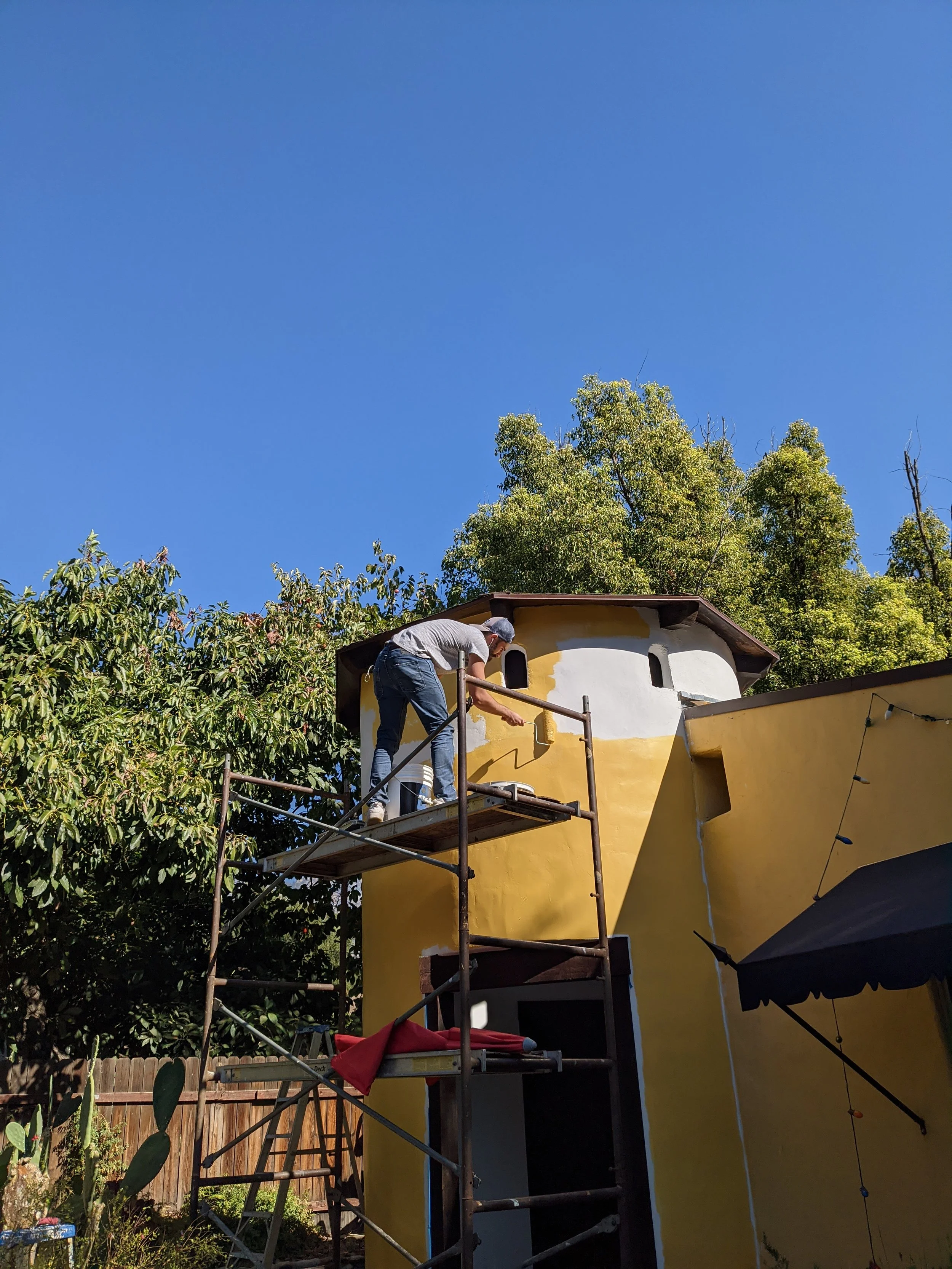 Person painting the exterior of a yellow and white house while standing on scaffolding under a blue sky with trees in the background.