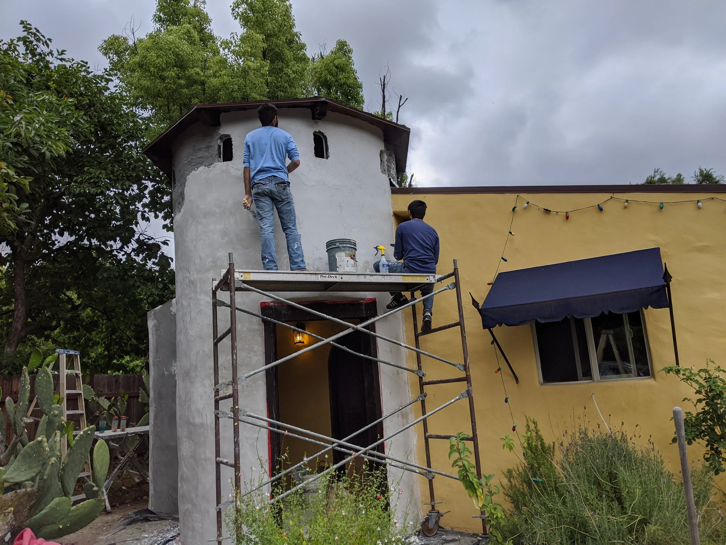 Two men working on the exterior of a building, one standing on scaffolding and the other sitting on it, with trees and a cloudy sky in the background.