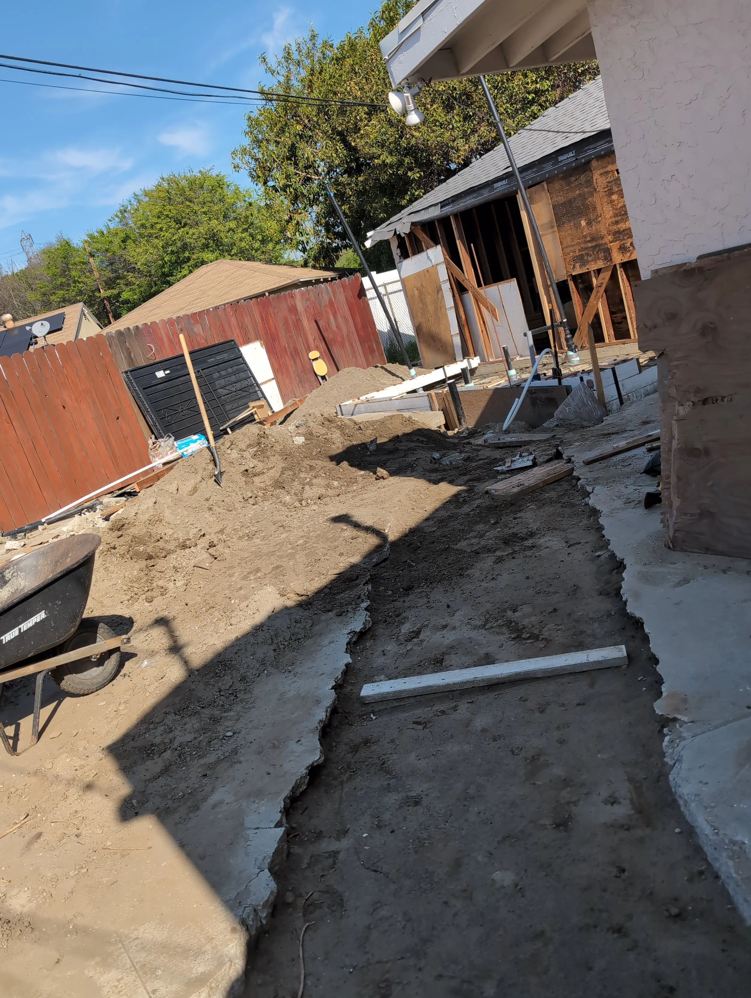 Construction site with dirt and concrete, partially built structure, wheelbarrow, and fencing in a backyard.