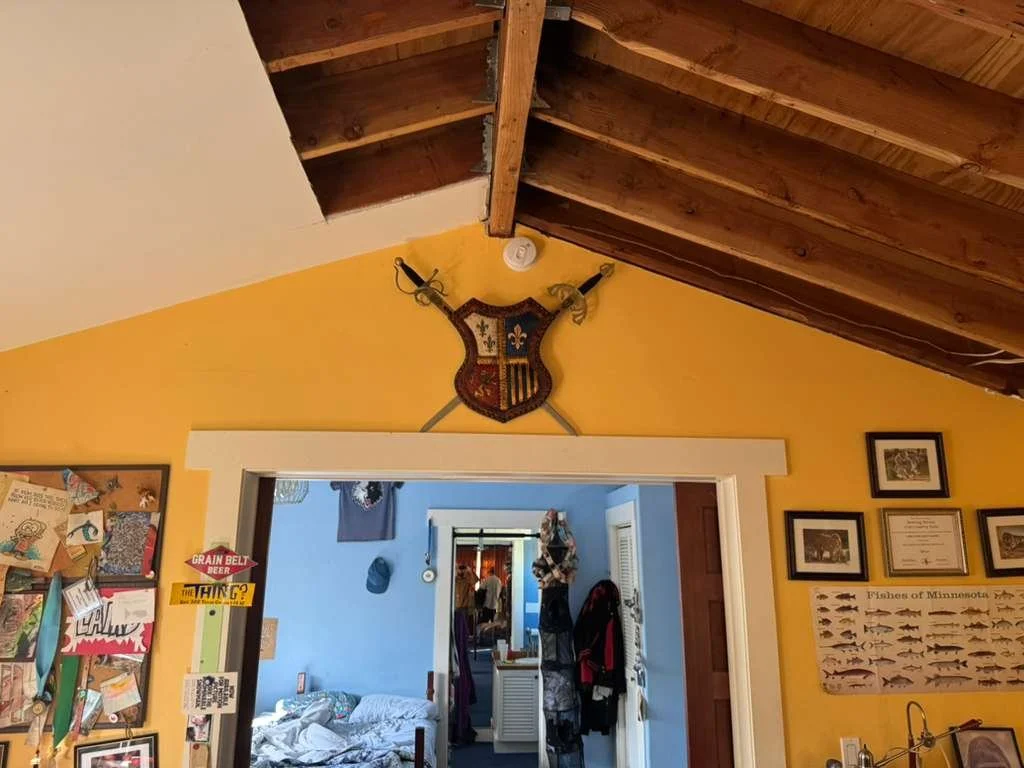 Interior of a room with a yellow wall and a wooden ceiling. A shield and swords are mounted above a doorway, leading to a blue-walled room with a bed and various items. The yellow wall has framed pictures and a poster of fish.