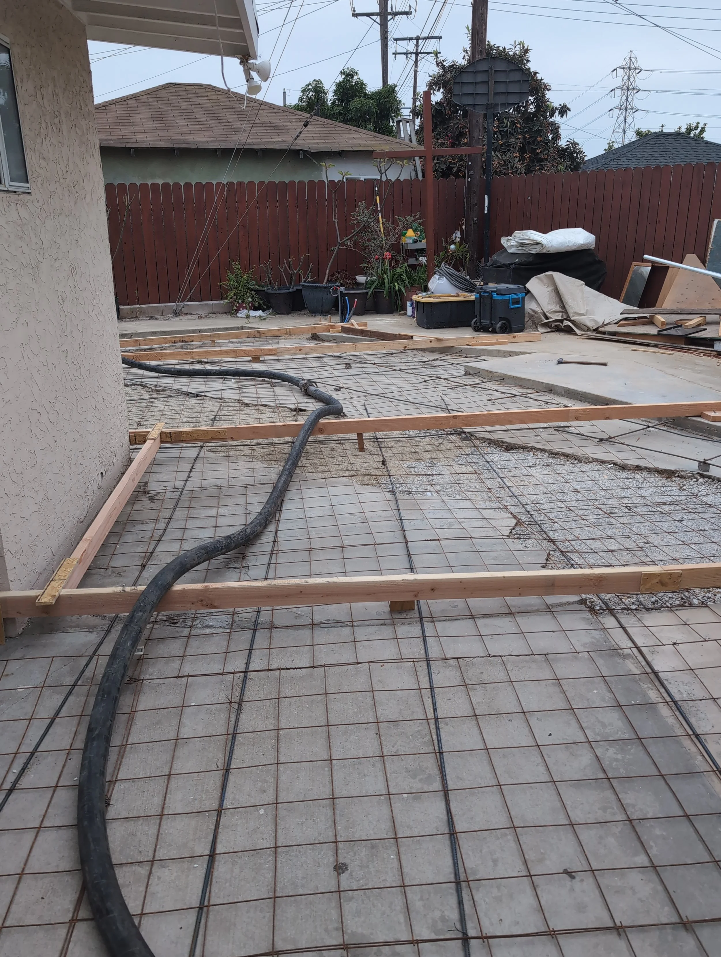 Construction site in a backyard with rebar and wooden framing for a concrete slab, black hose on the ground, and potted plants along a wooden fence in the background.