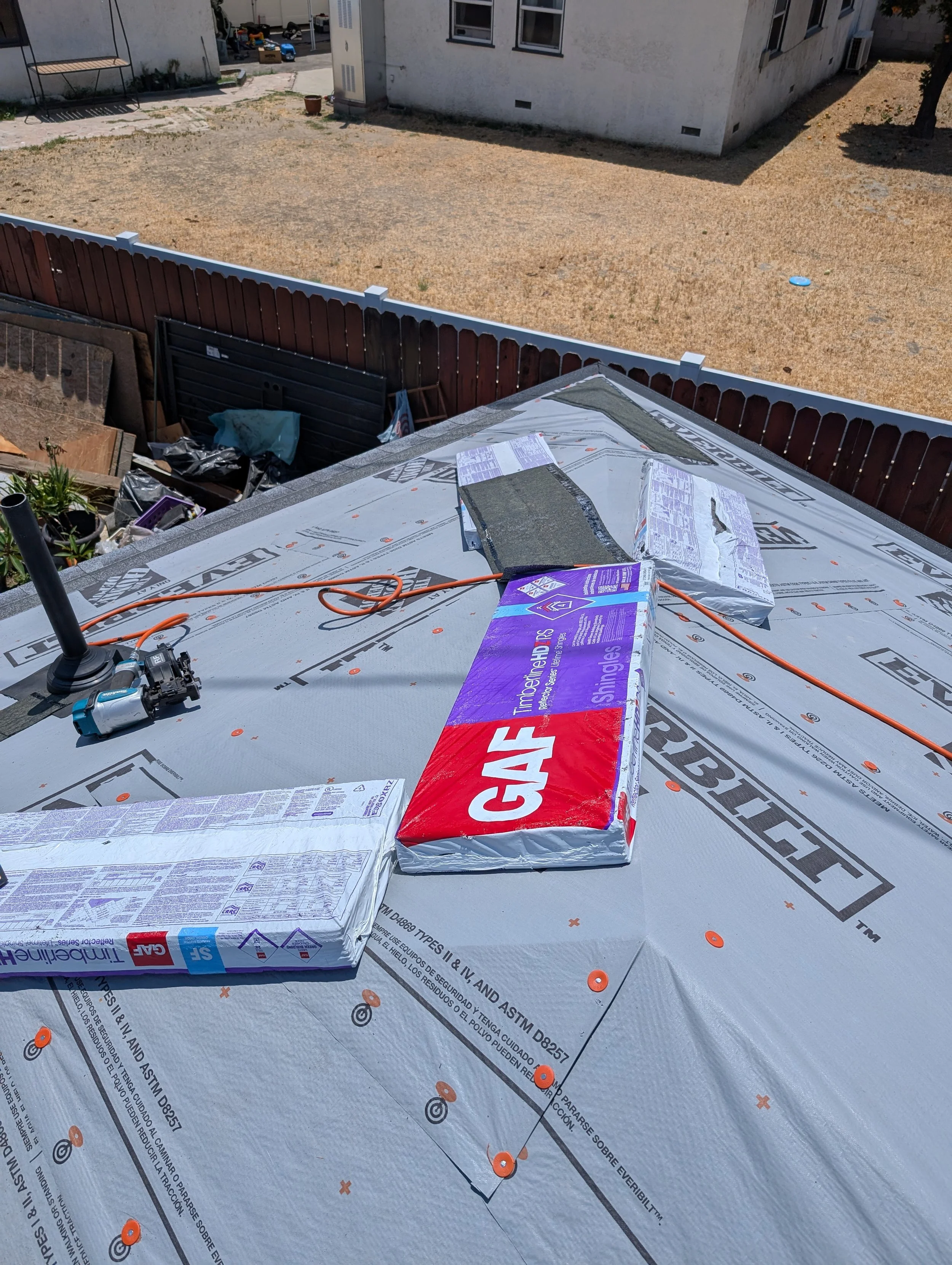 Roof under construction with insulation materials, tools, and orange cords, with a backyard and neighboring house in the background.