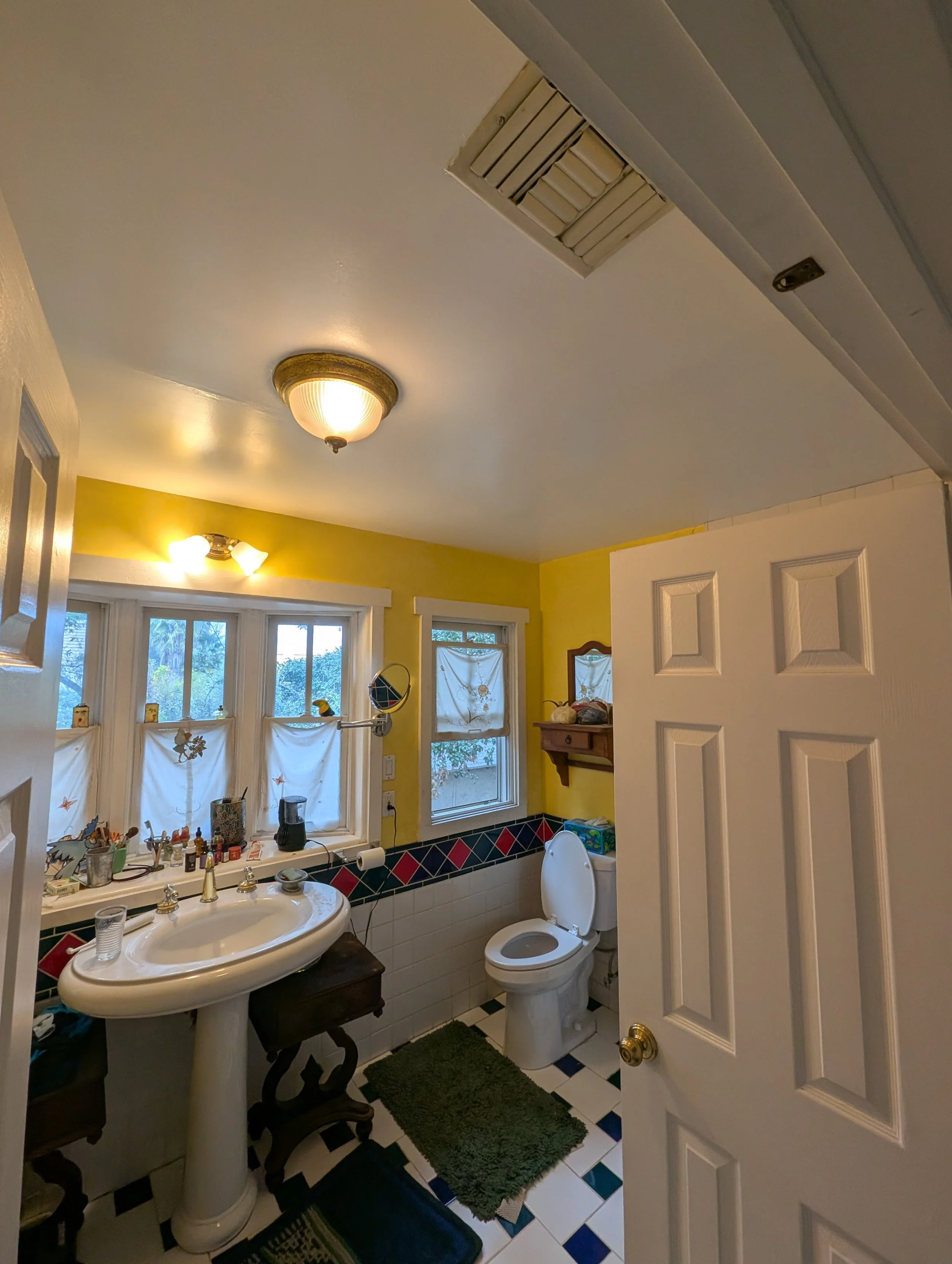 A small bathroom with a yellow wall, a pedestal sink, a toilet, three windows with white curtains, and various toiletries and decorations. There are green and black rugs on the black and white checkered floor, and a ceiling light fixture.