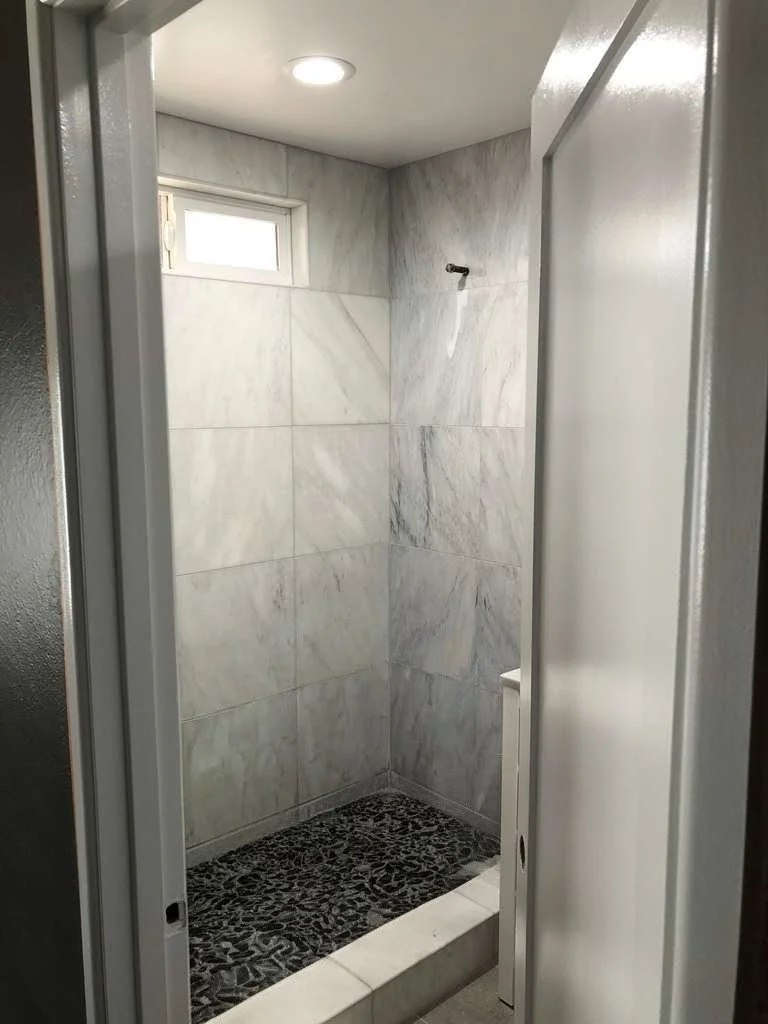 A small, minimalist shower stall with marble tiles on the walls, a pebble stone shower floor, a small window, and a ceiling light.