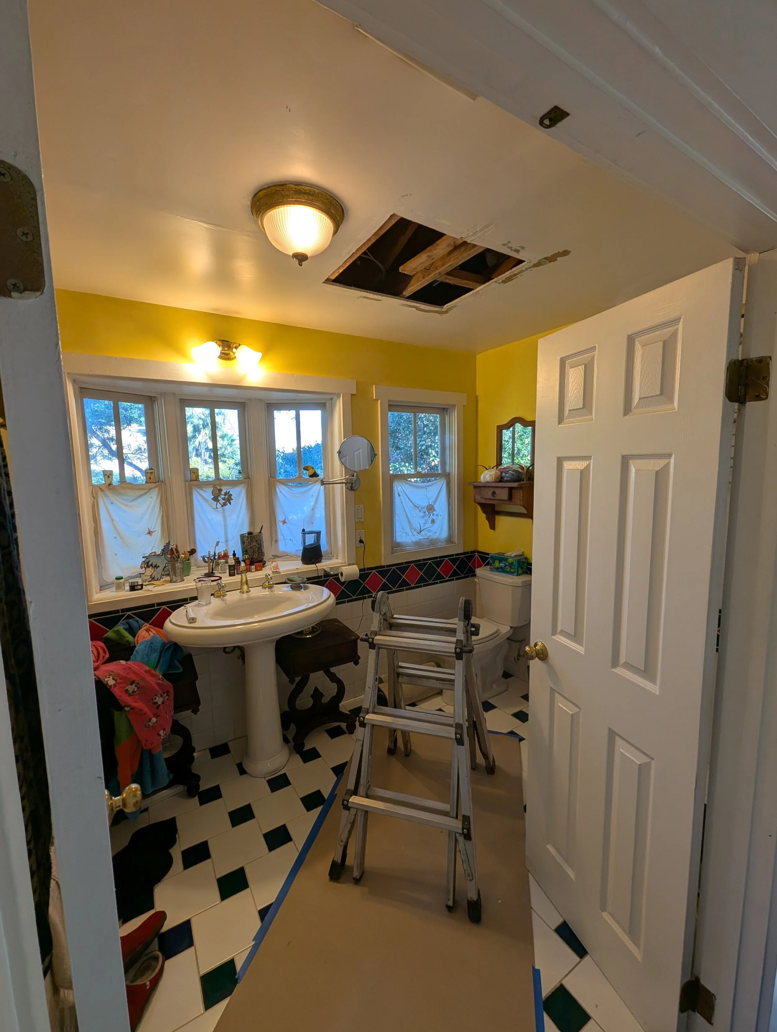 A cluttered bathroom with a yellow wall, black and white checkered tile floor, and a ceiling with a hole revealing wooden beams. It has a sink with various items on the windowsill, a mounted mirror, a toilet, and a small wooden shelf. A stepladder is