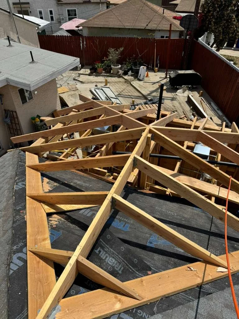 Construction site with wooden framework for a roof or floor being built, surrounded by a backyard with potted plants, a fence, and neighboring houses.