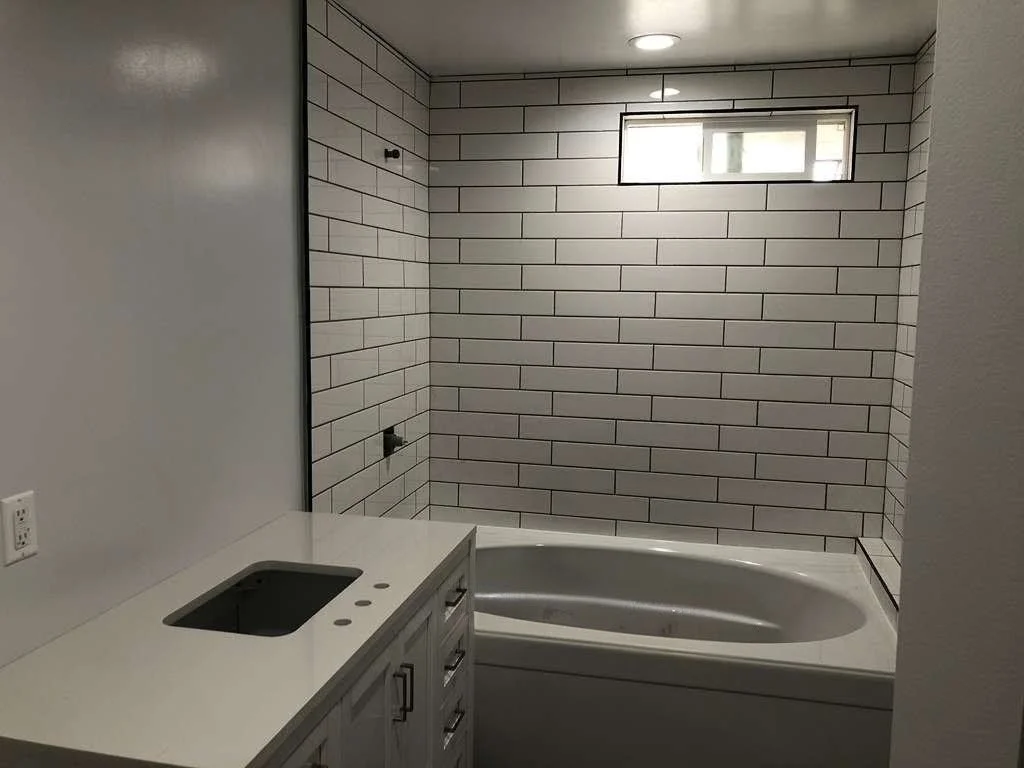Bathroom with white countertop, small black sink, and bathtub with tiled wall in the background, a small window, and a ceiling light.