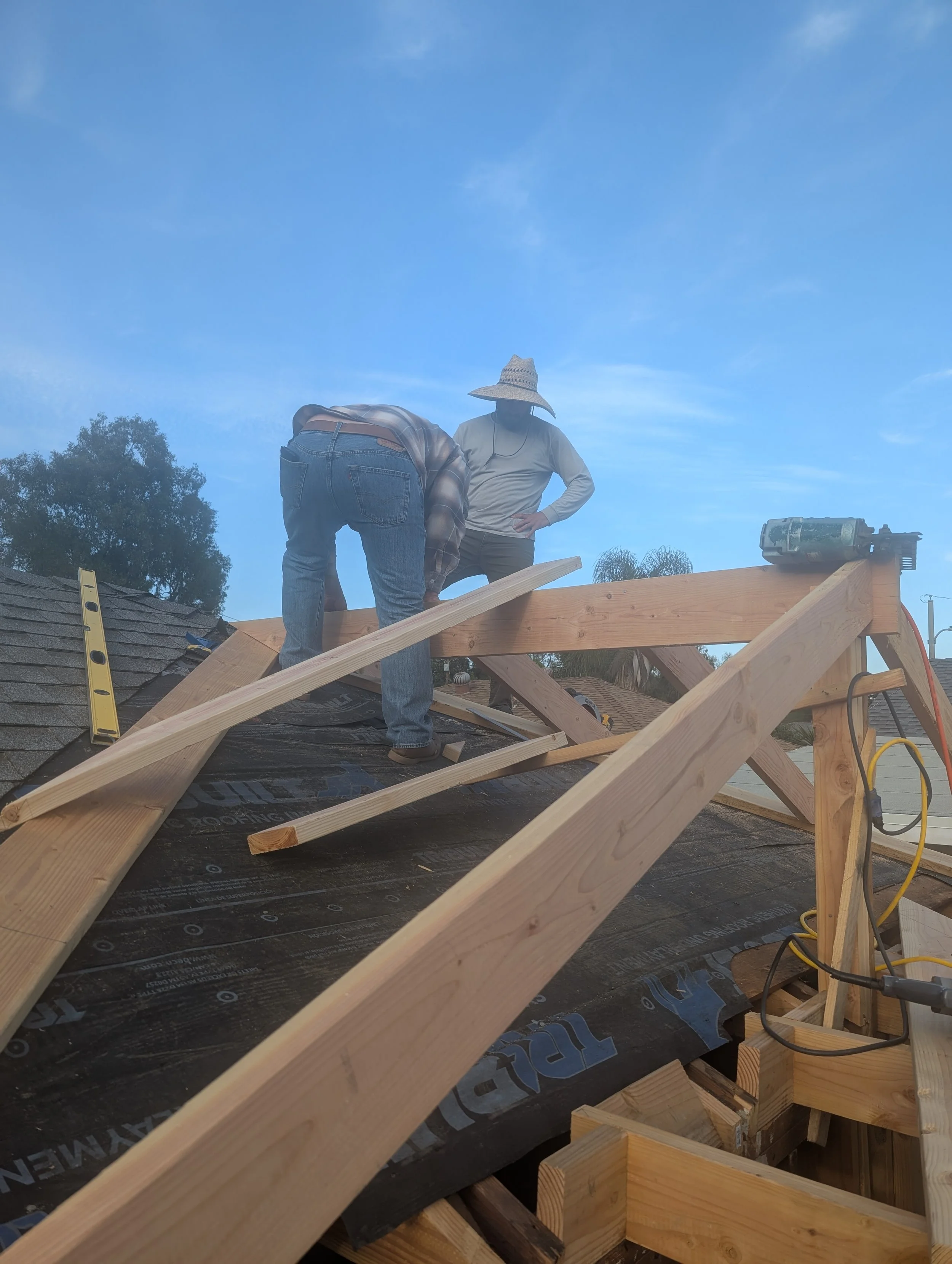 Two workers constructing the roof of a building with wooden beams and black roofing material under a blue sky with some clouds.