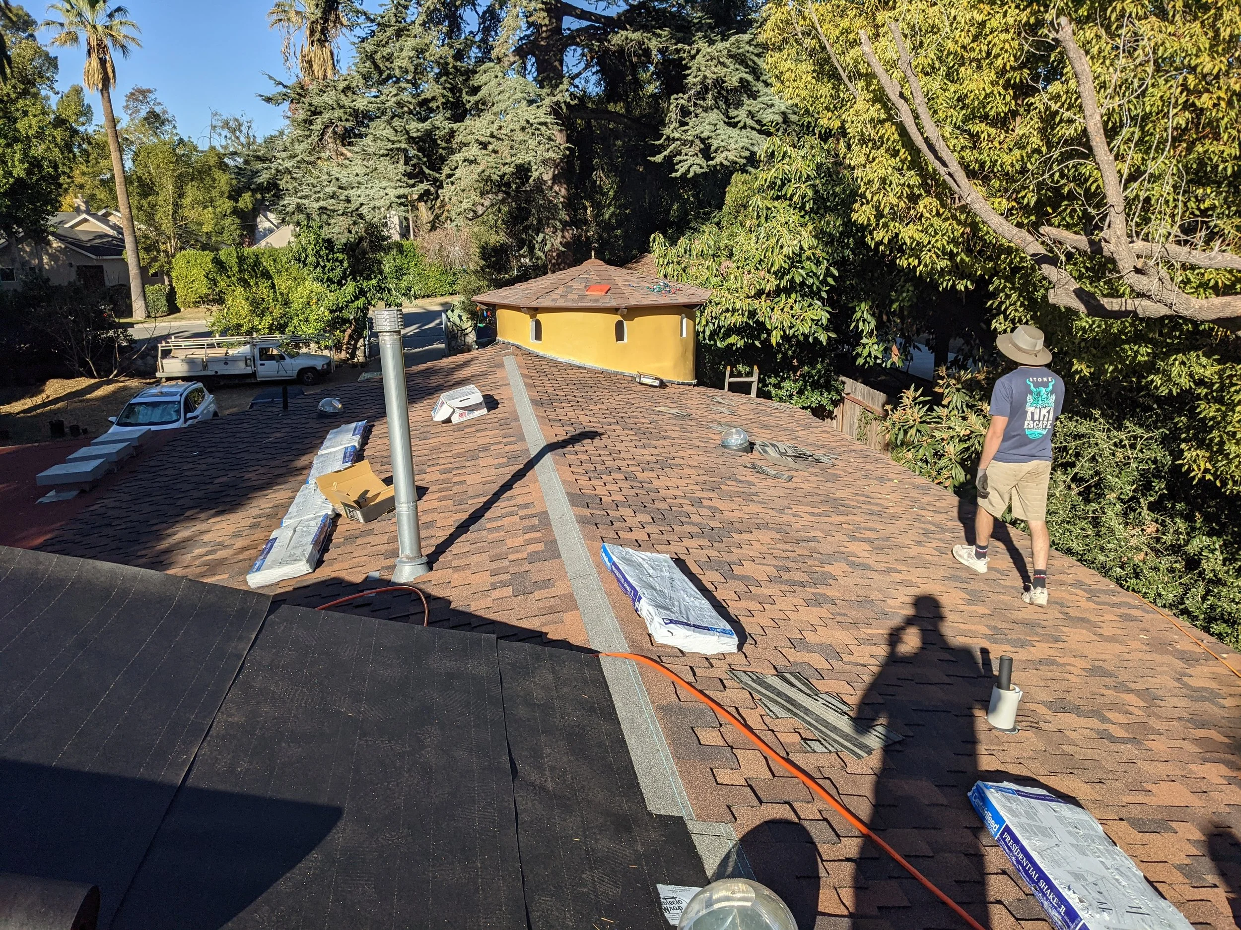A person wearing a wide-brimmed hat, a blue T-shirt, shorts, and sneakers standing on a rooftop under a tree, with building materials and tools scattered around.