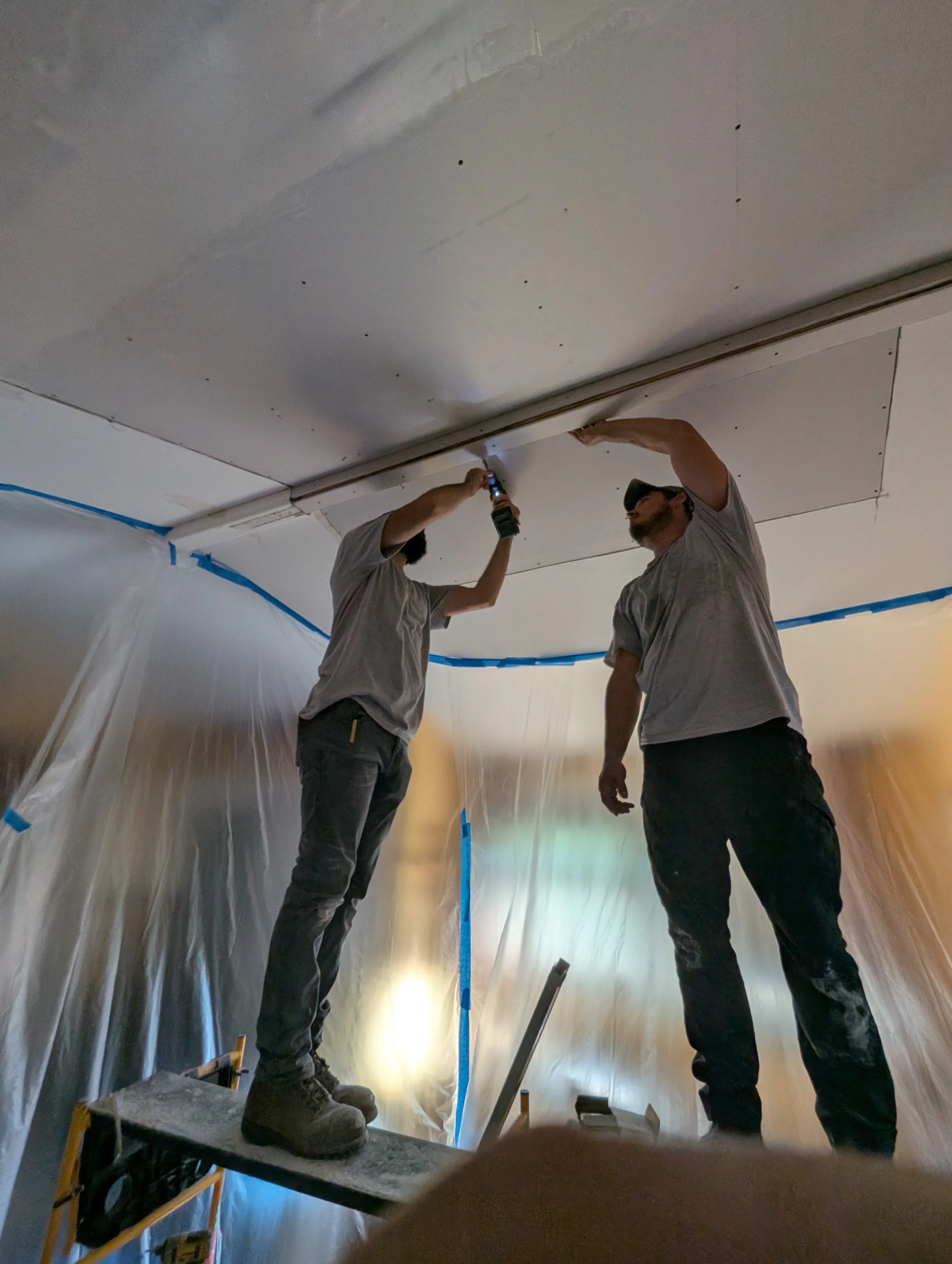 Two workers installing a ceiling panel, one with a drill, in a room covered with plastic sheeting and painter's tape.
