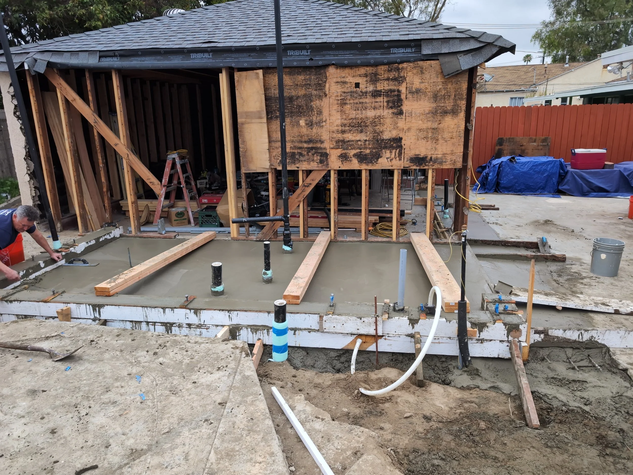 Construction site with a concrete slab foundation being poured in front of a small wooden structure under renovation, with tools and construction materials visible.