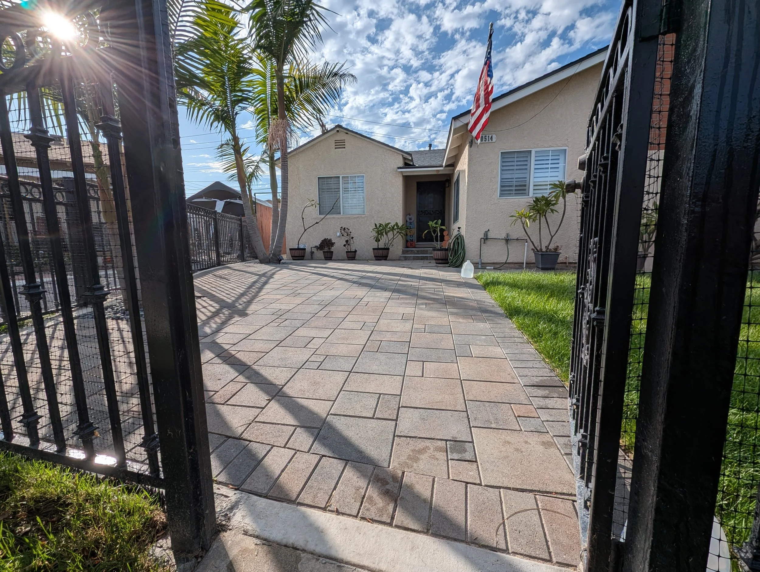 View of a house with a small front yard, potted plants, and a paved walkway, seen through an open black wrought iron gate. The house has beige walls, a front door, windows with shutters, an American flag, and a blue sky with scattered clouds.