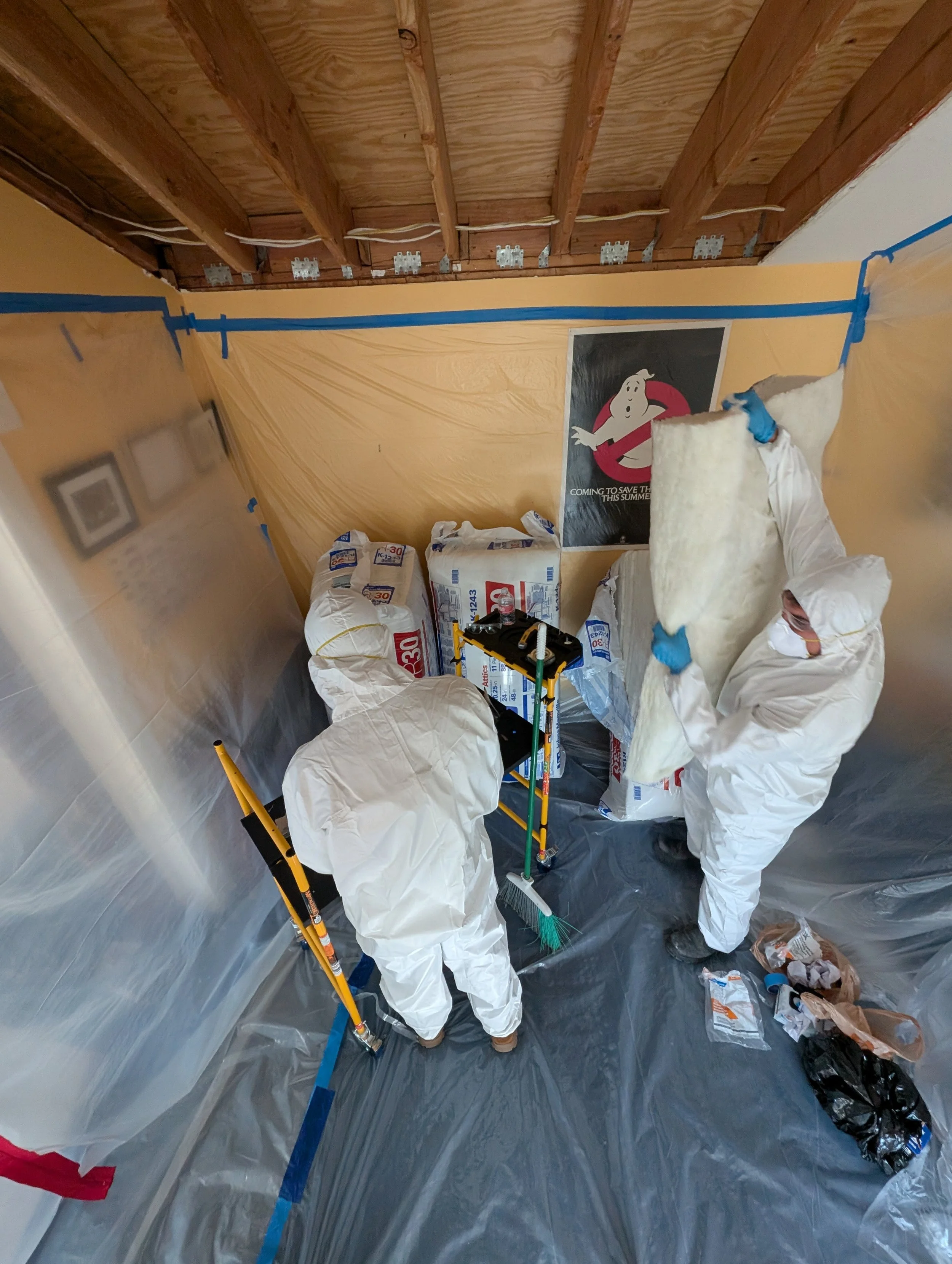 Two workers in white protective suits installing insulation into a wall cavity that is covered with plastic sheeting in a room. The workers are using tools and wearing blue gloves. The room has exposed wooden ceiling beams and a poster of the Ghostbu