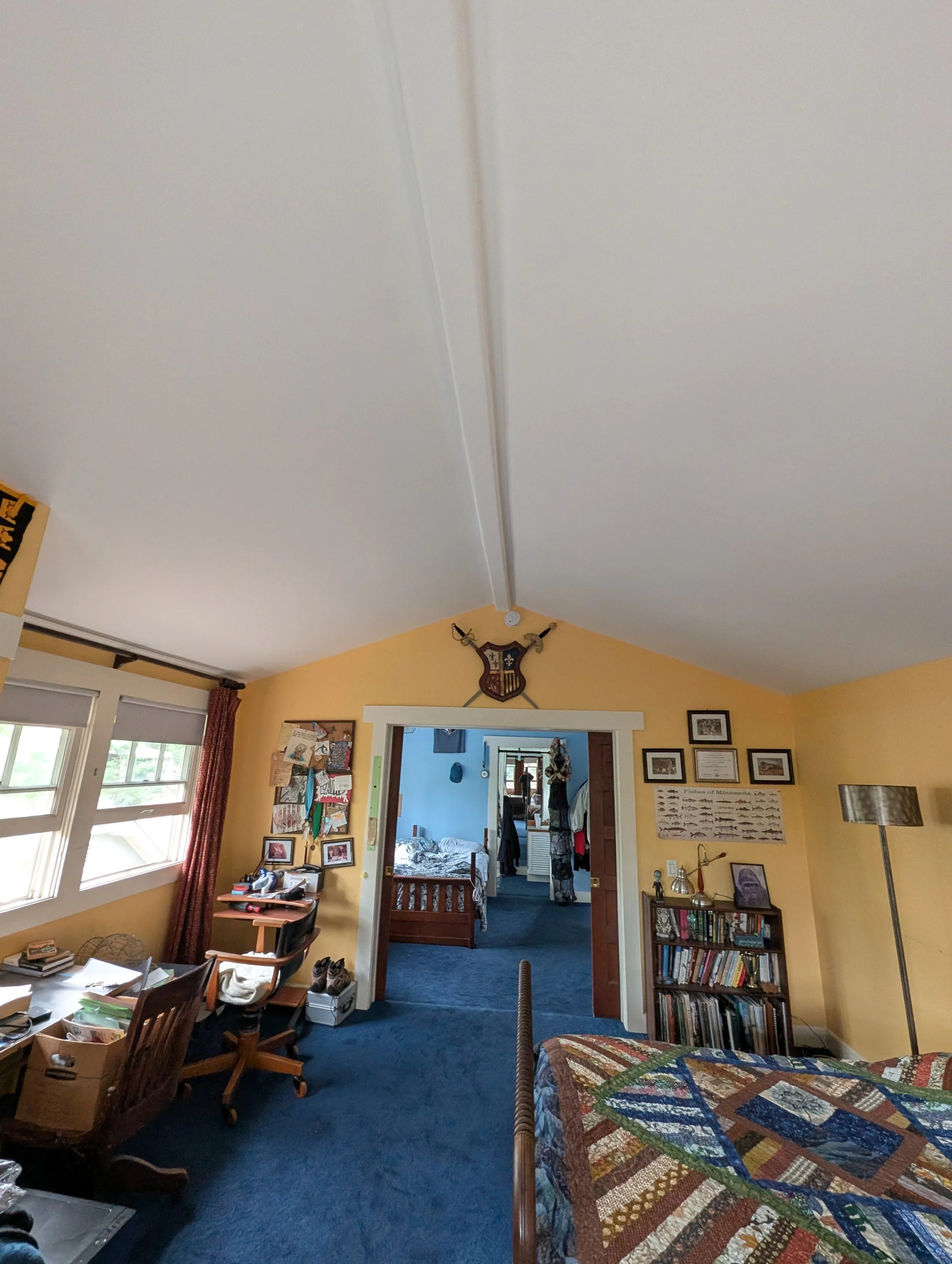 A view of a cozy bedroom with yellow walls, a patchwork quilt on the bed, a bookshelf filled with books, a tall lamp, and framed pictures on the wall. There is a doorway leading to another room with a bed and a person visible in the reflection of a m