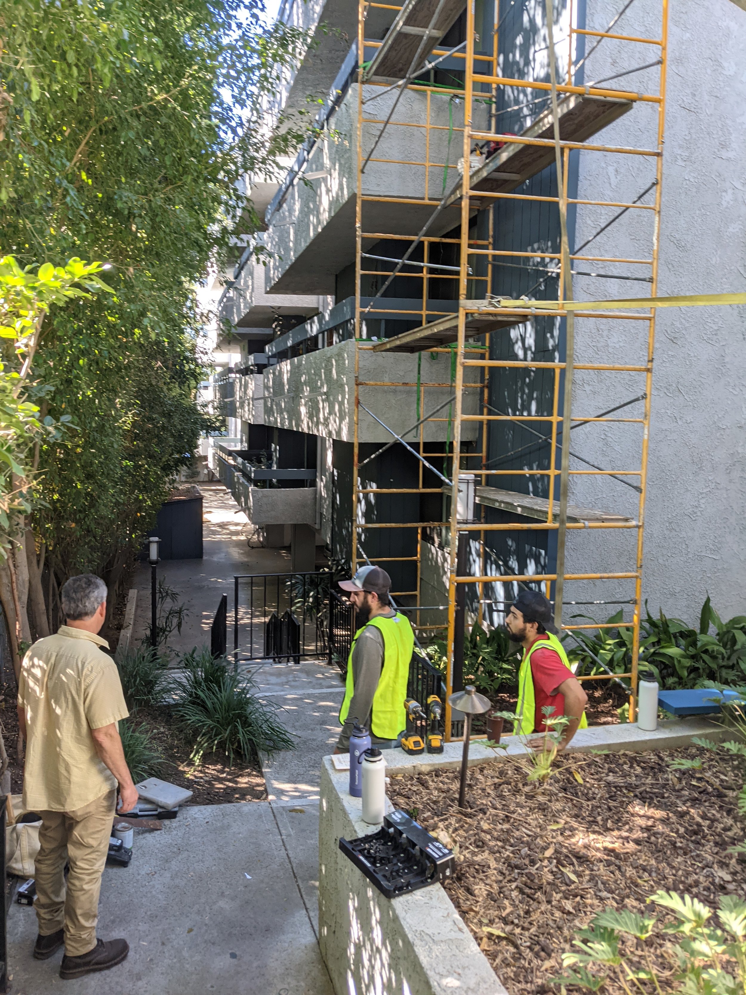 Three men working on a building with scaffolding; two wearing yellow safety vests and one in a tan shirt, standing near construction tools and supplies outside.
