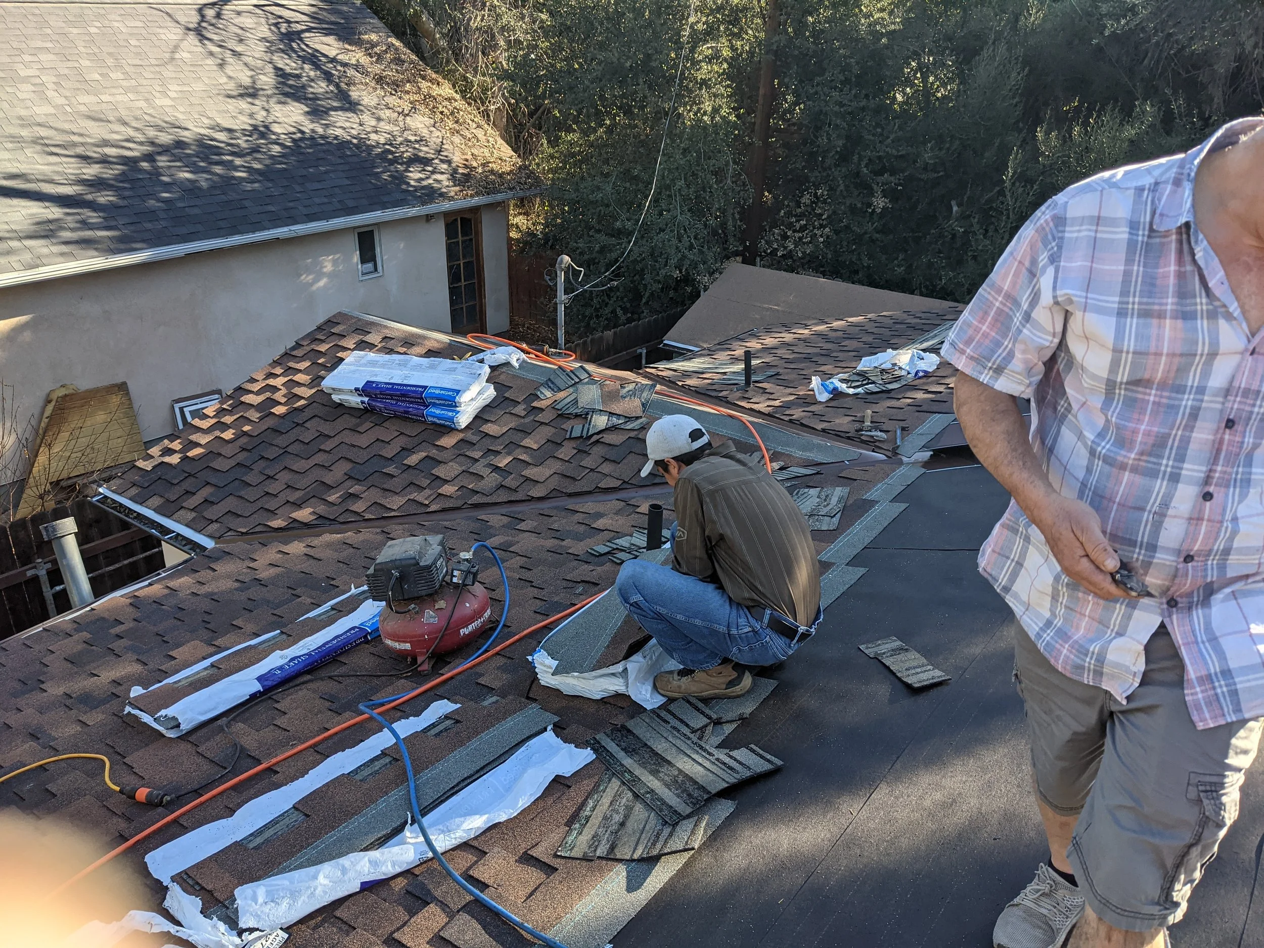 Two men installing or repairing a roof with shingles on a house, with roofing materials and tools around.