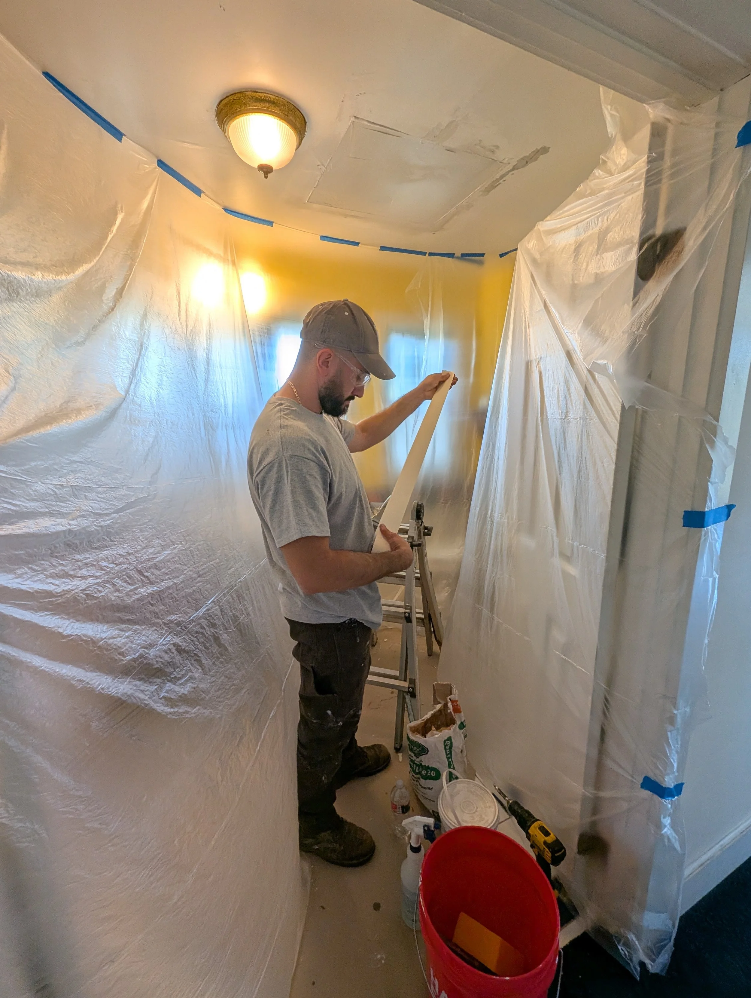 A man working on a home improvement project inside a small room with plastic sheeting taped to the walls, wearing a gray T-shirt, black pants, a gray cap, and safety glasses, using a tool on a long piece of material.