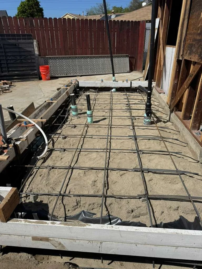 Construction site with rebar grid, plumbing pipes, and wooden framing in a backyard.