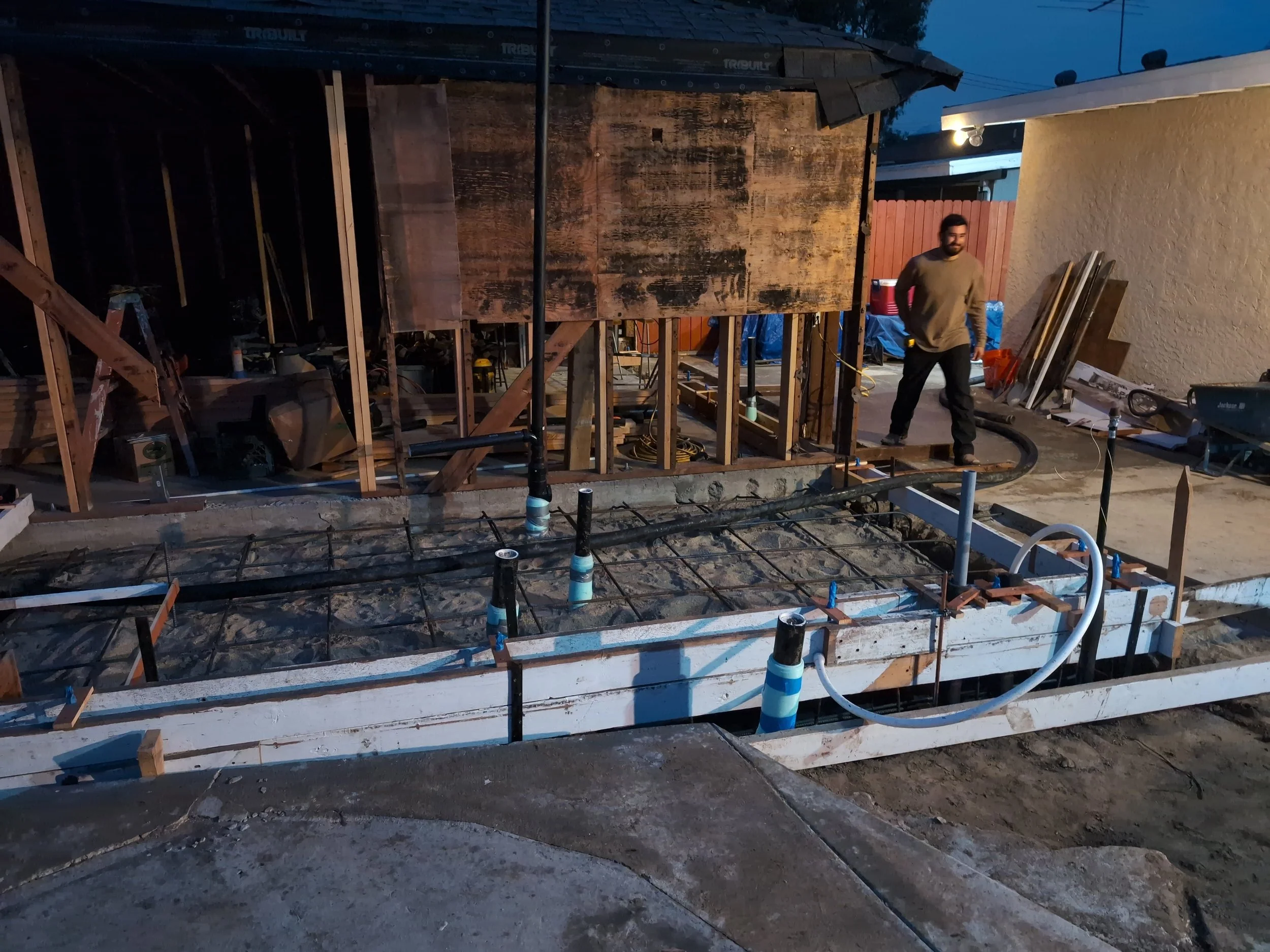 Construction worker standing on a concrete slab with exposed rebar, framing and plumbing pipes, during early building stage outside of a house at dusk.