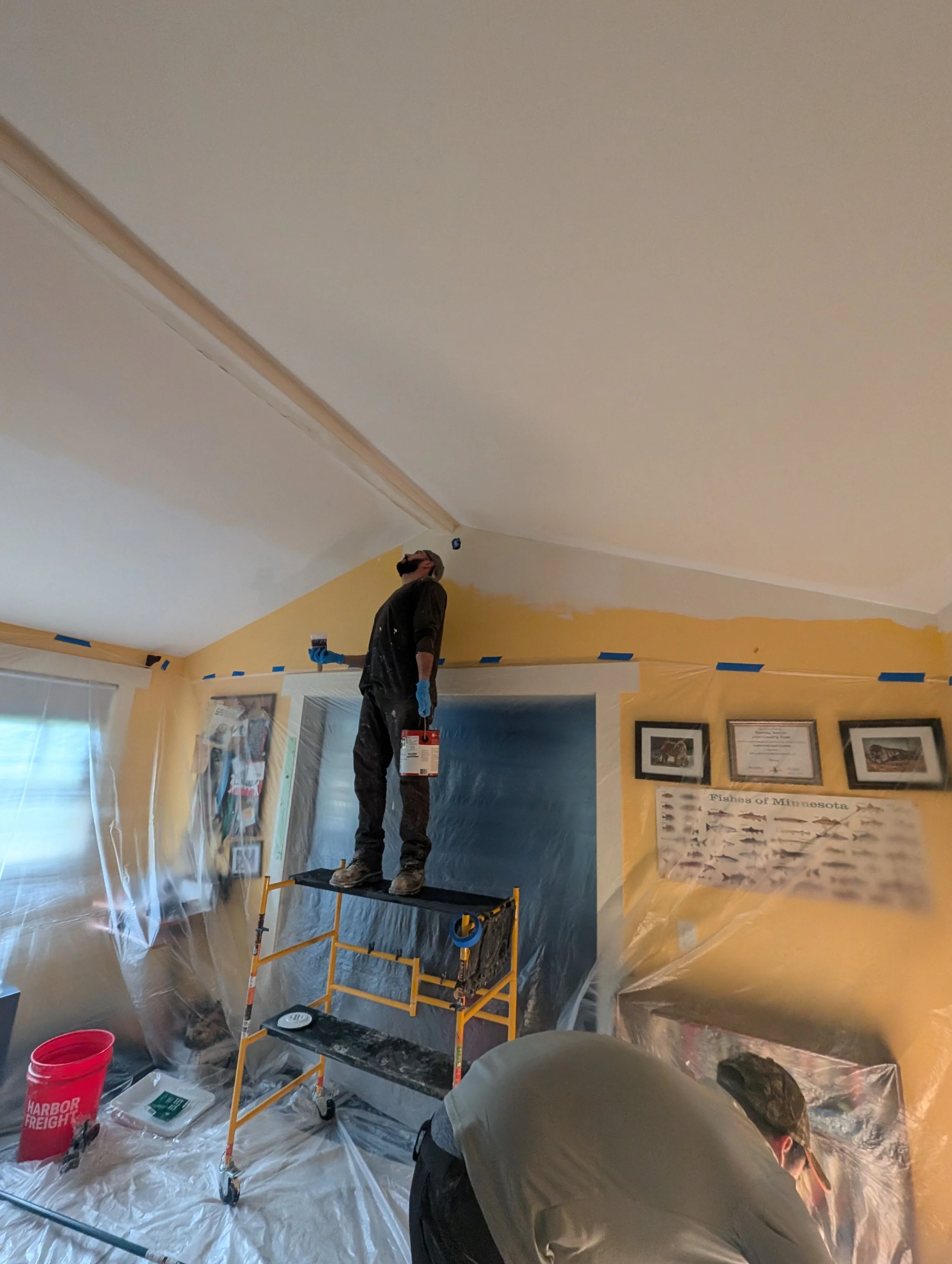 Person standing on scaffold painting a room ceiling white while another person works below, with protective plastic covering furniture and walls