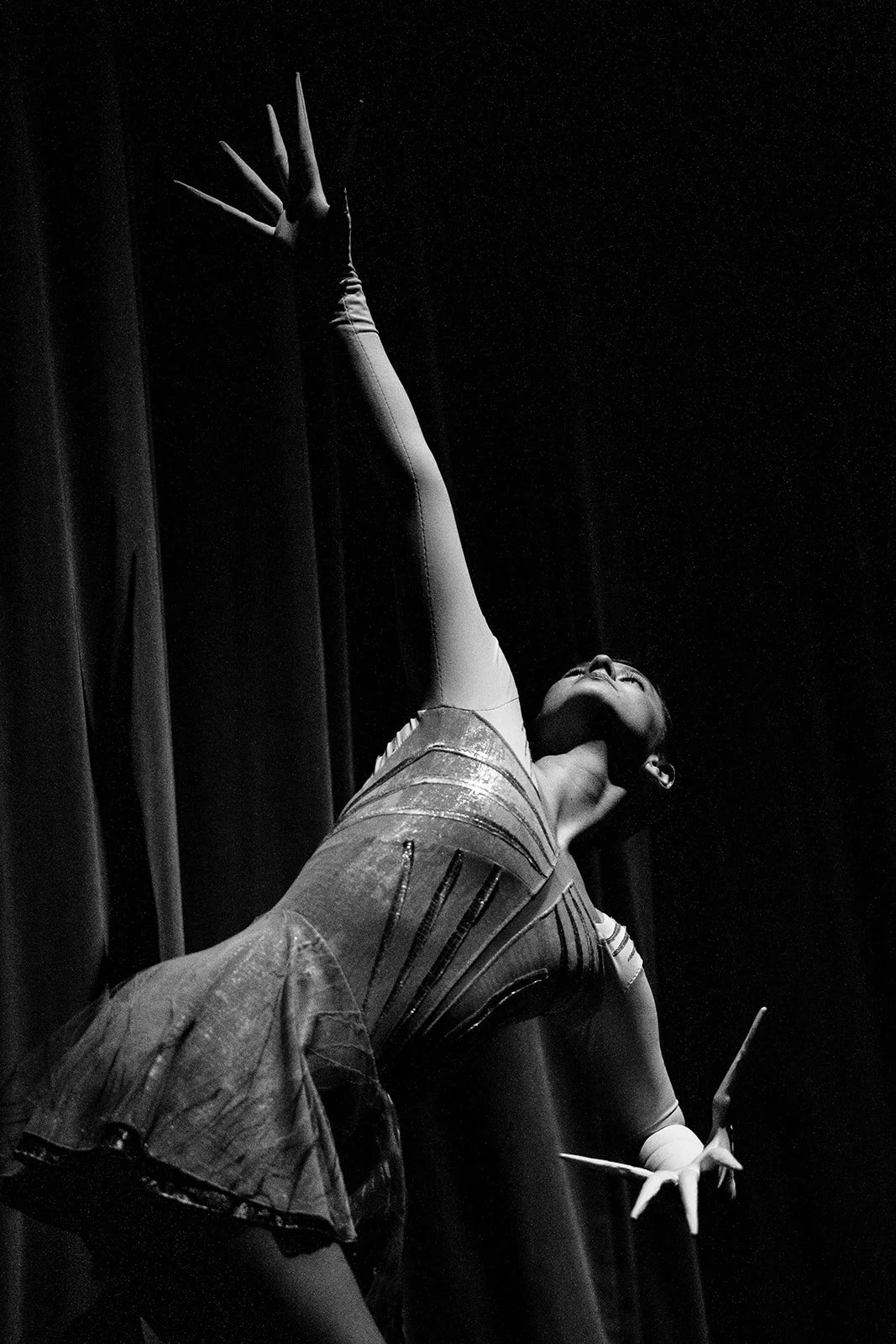 Black and white photograph of a ballerina on stage, stretching her arm upward with a dramatic pose, wearing a tutu and ballet shoes, with a dark curtain background.