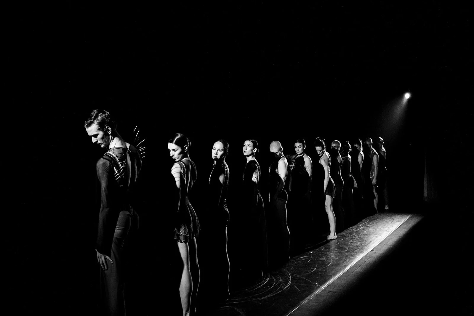 A line of models stands on a runway during a fashion show, illuminated by focused lighting in a dark setting.