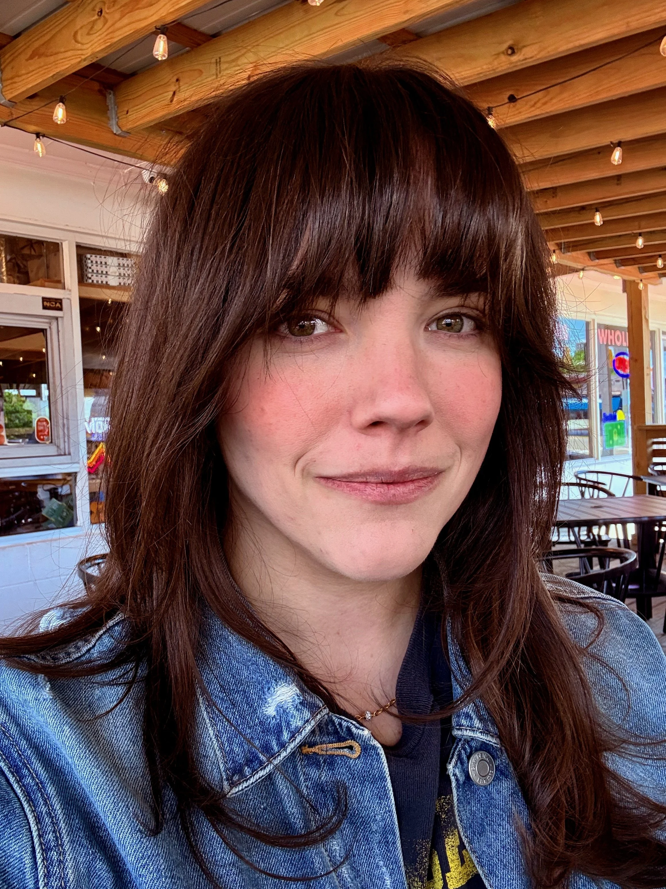 Close-up of a woman with long, dark brown hair and hazel eyes, smiling slightly, wearing a denim jacket and a necklace, sitting outside in a restaurant with wooden beams and string lights overhead.