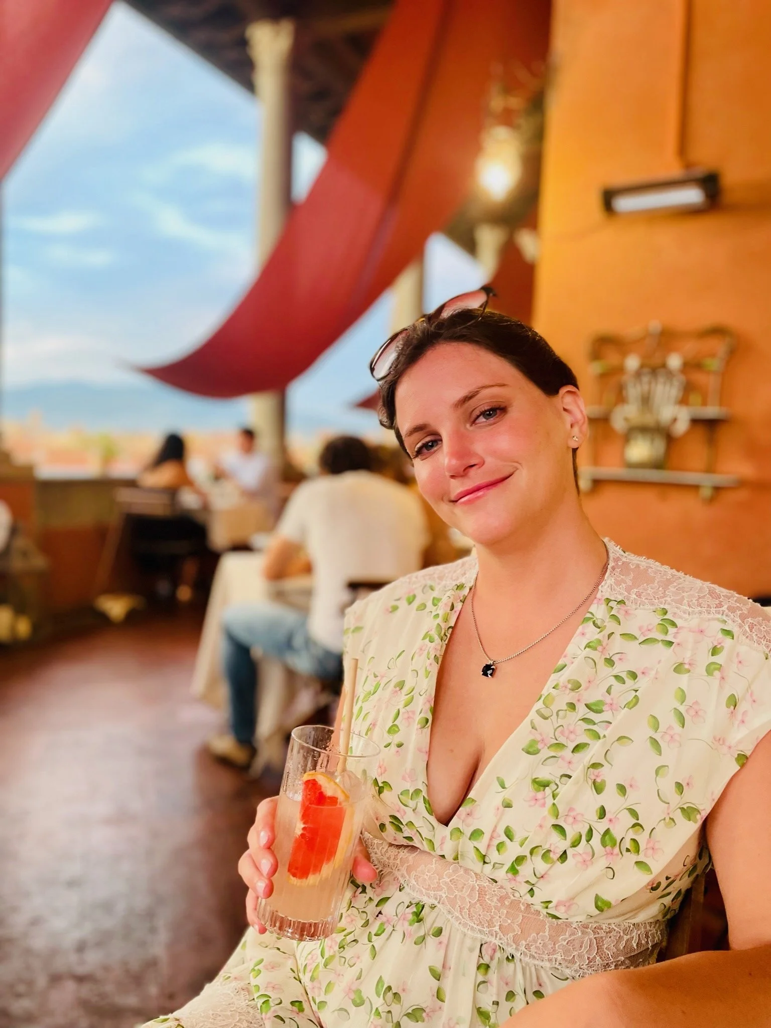 A woman with short dark hair and earrings sitting at an outdoor restaurant, holding a glass with a pink cocktail and strawberry slice, smiling at the camera, with a blurred background of other diners and a colorful awning.