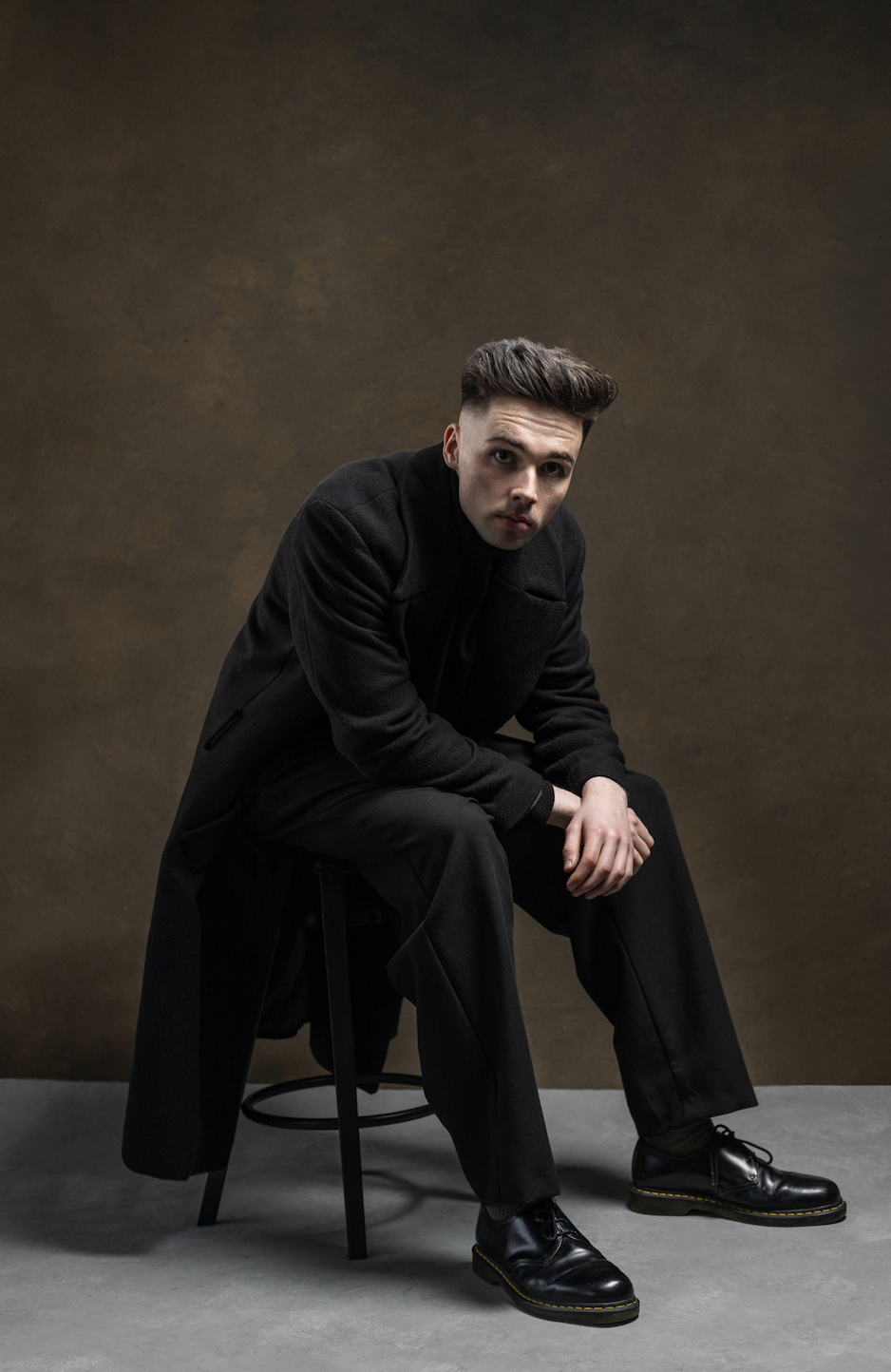 A young man with dark hair styled up, dressed in black, sitting on a stool with a dark brown background.