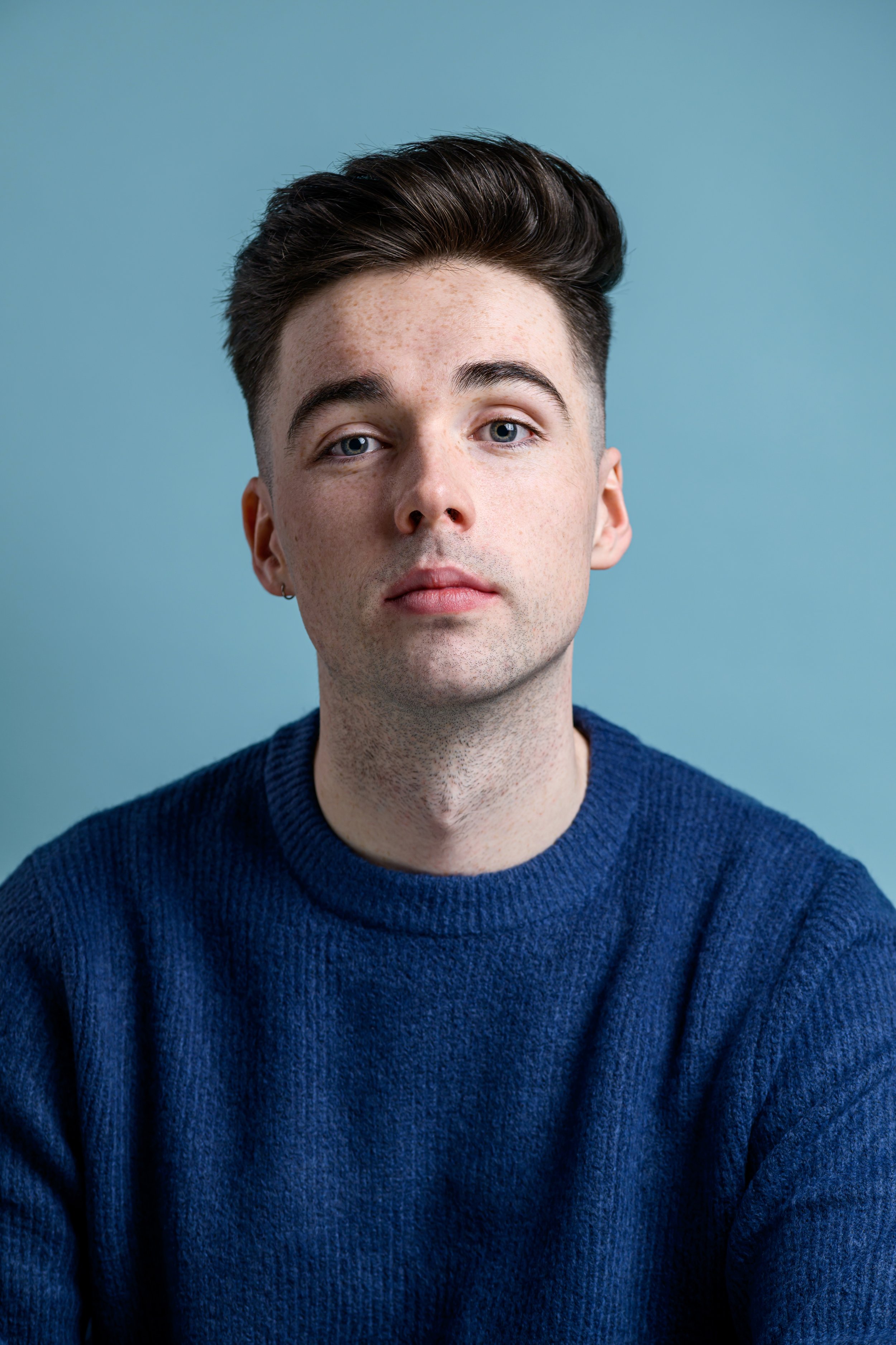 A young man with short dark hair, blue eyes, and freckles, wearing a navy blue sweater, stands against a light blue background.